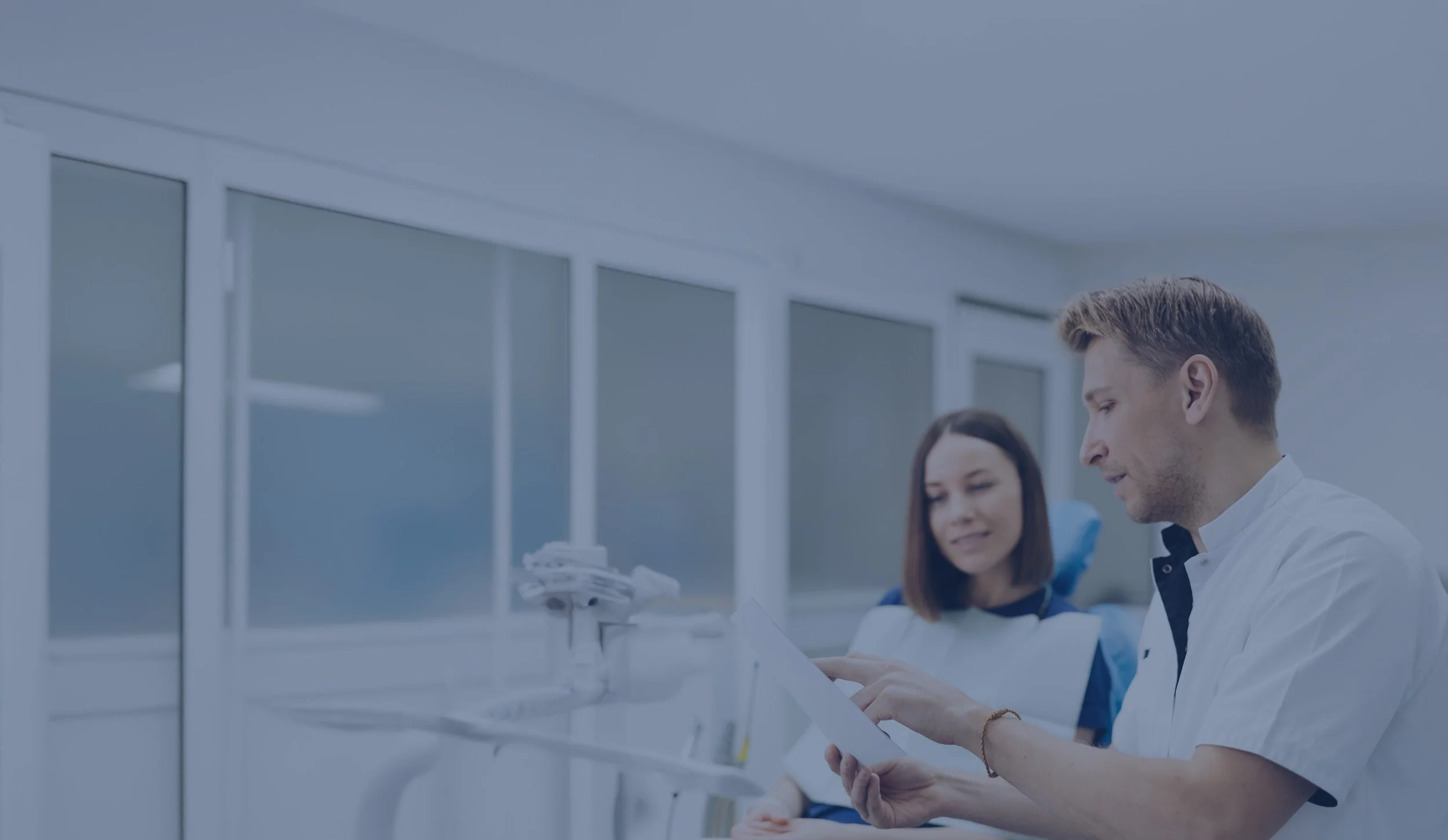 A male and female healthcare professionals sitting together in a medical facility, looking at a tablet and a file, with large windows in the background.
