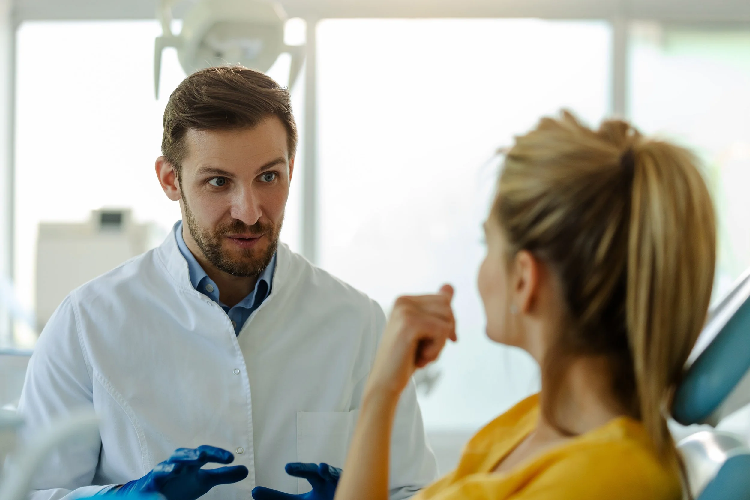 A male doctor or dentist speaking to a female patient in a clinical setting. The woman is seated in a dental chair, and the man appears to be explaining something or providing reassurance.