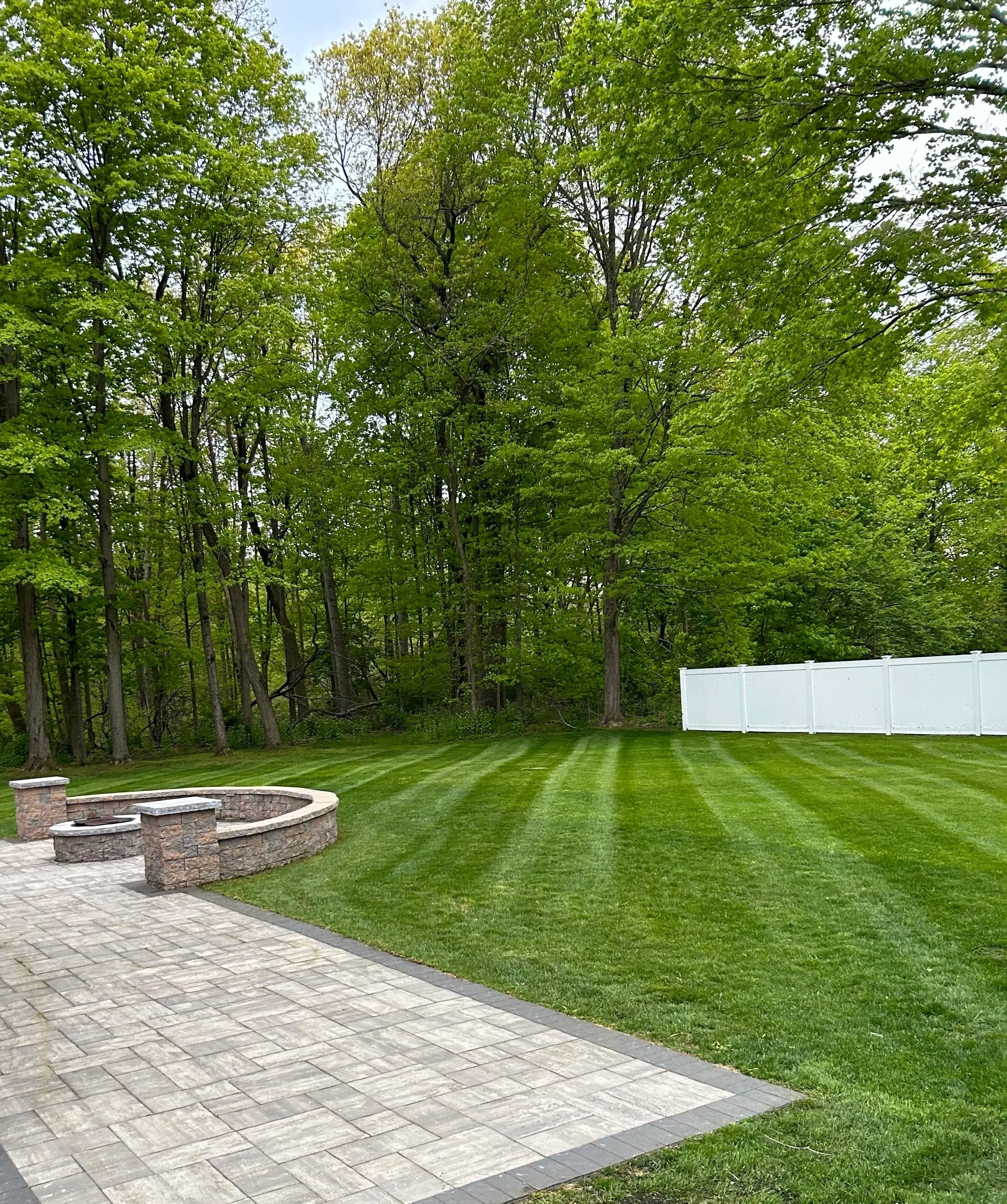 Backyard with green grass, a stone patio with firepit, and a wooded area in the background.