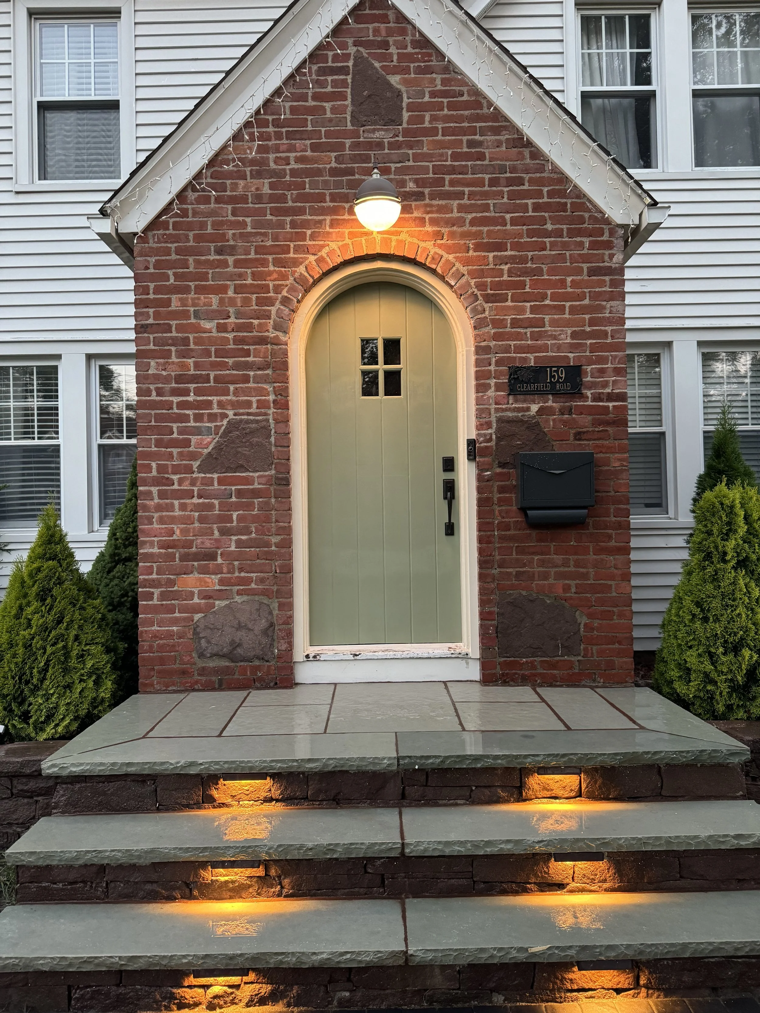 The image shows the front entrance of a house with a brick facade, featuring a light green door with a small window, a black mailbox, and an address plaque, illuminated by exterior lighting. There are steps with lighting leading up to the door, flanked by small bushes.