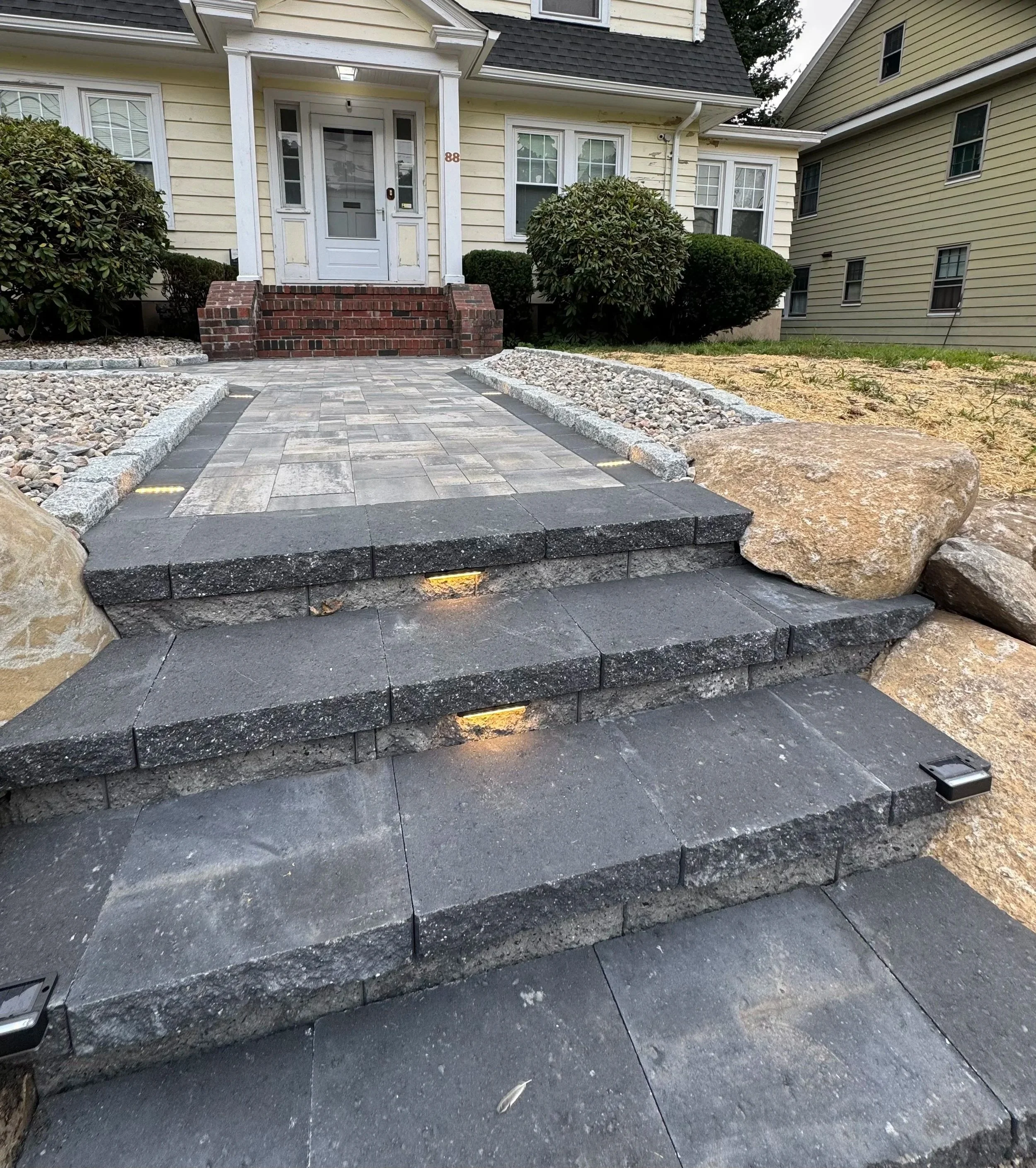 Stone pathway leading to front porch with stairs, surrounded by rocks and landscaping, residential house in background.