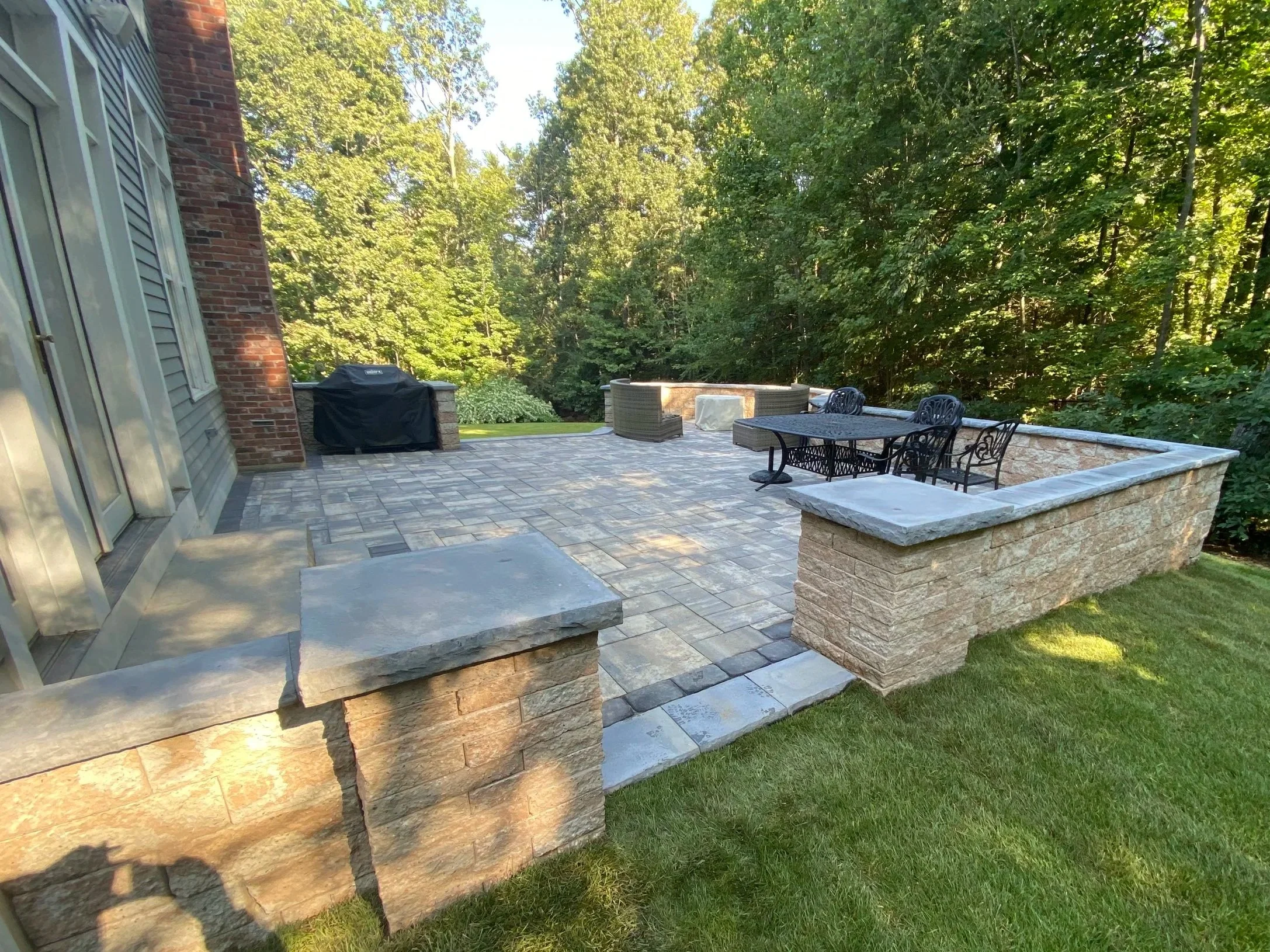 A spacious outdoor patio with stone brick walls, a tiled floor, and a wrought iron table with chairs surrounded by a wooded area.