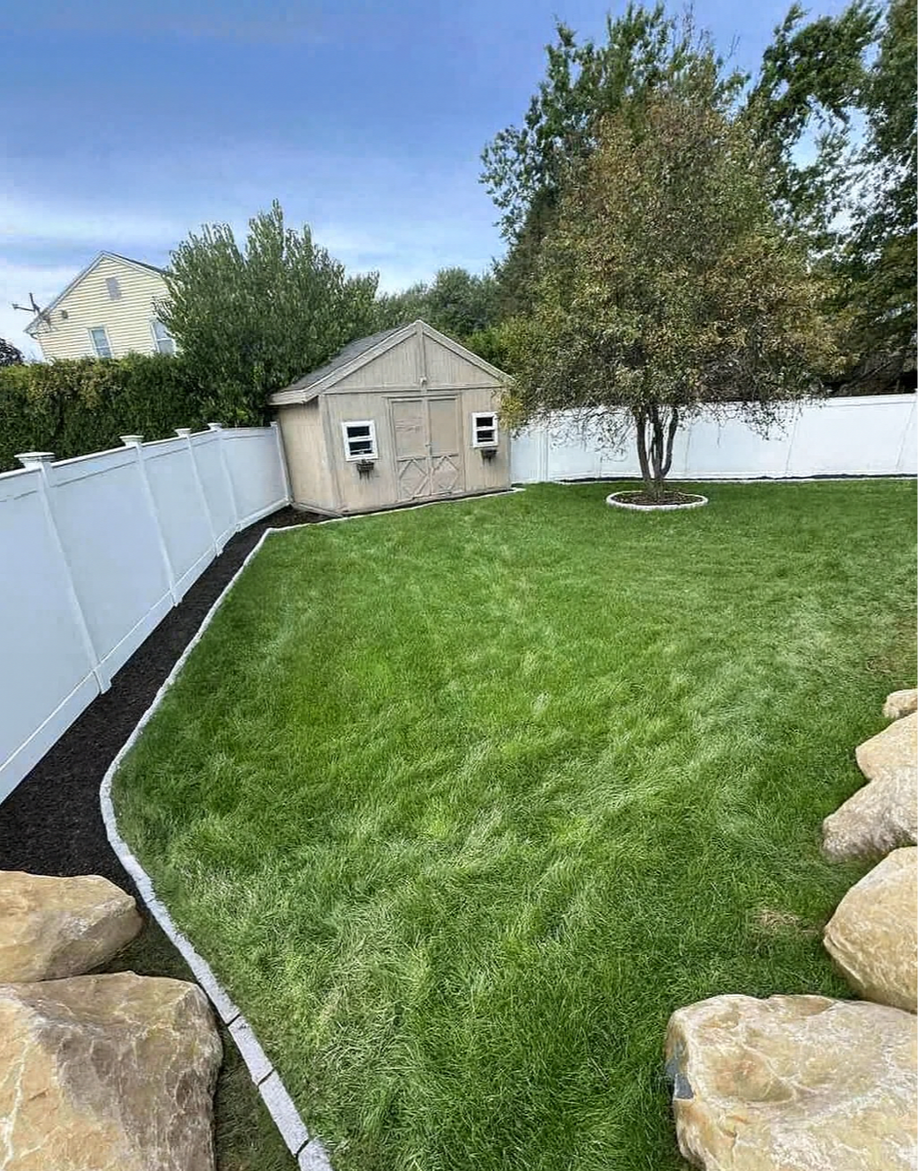 A well-maintained backyard with green grass, a white fence, a small beige shed, and a tree with a circular flower bed. Large rocks are in the foreground.
