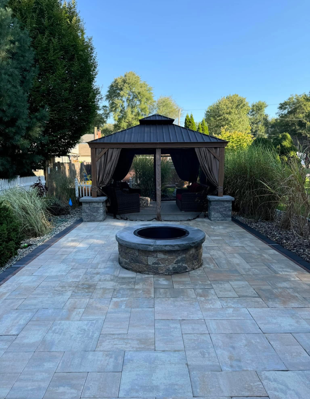 A backyard patio with a stone fire pit in front and a gazebo with curtains behind.