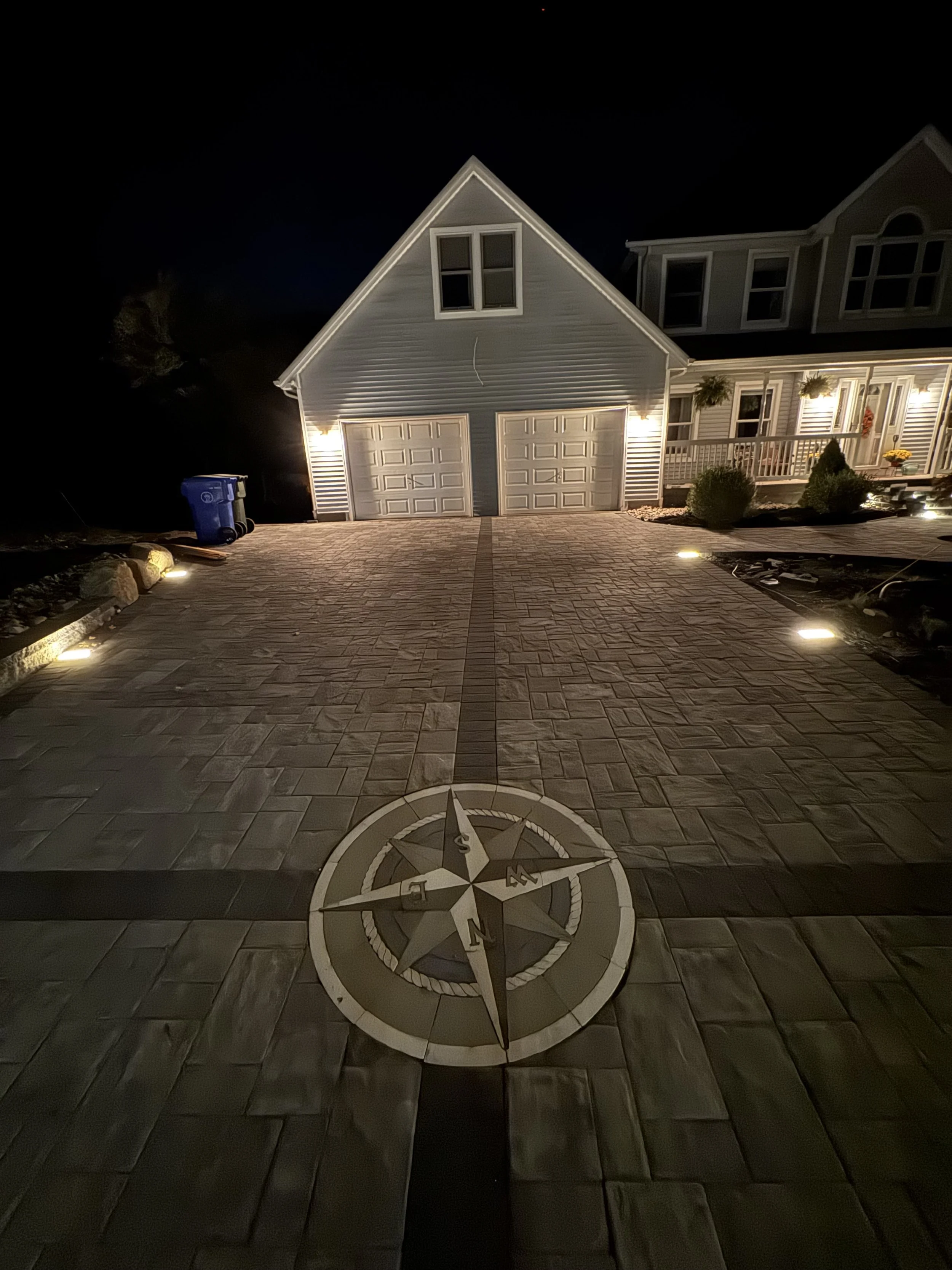 Night view of a residential driveway with a compass design, leading up to a garage and house, illuminated by outdoor lights.
