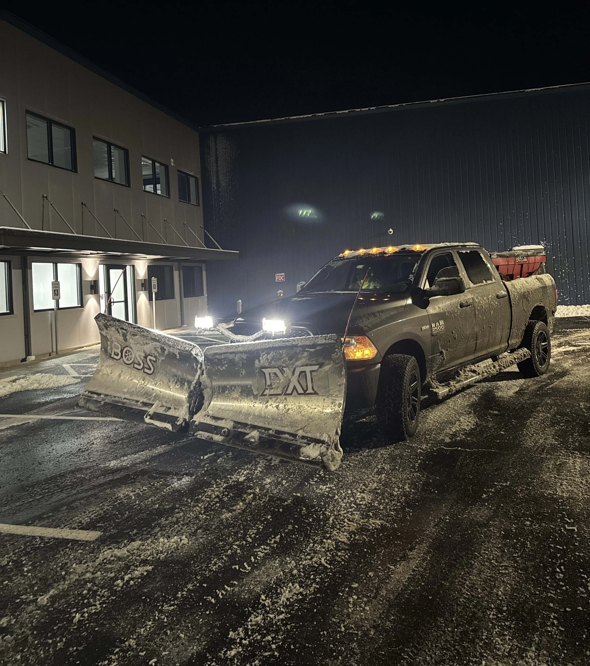 A black pickup truck with a snowplow attached to the front, parked outside a building at night in snowy conditions.