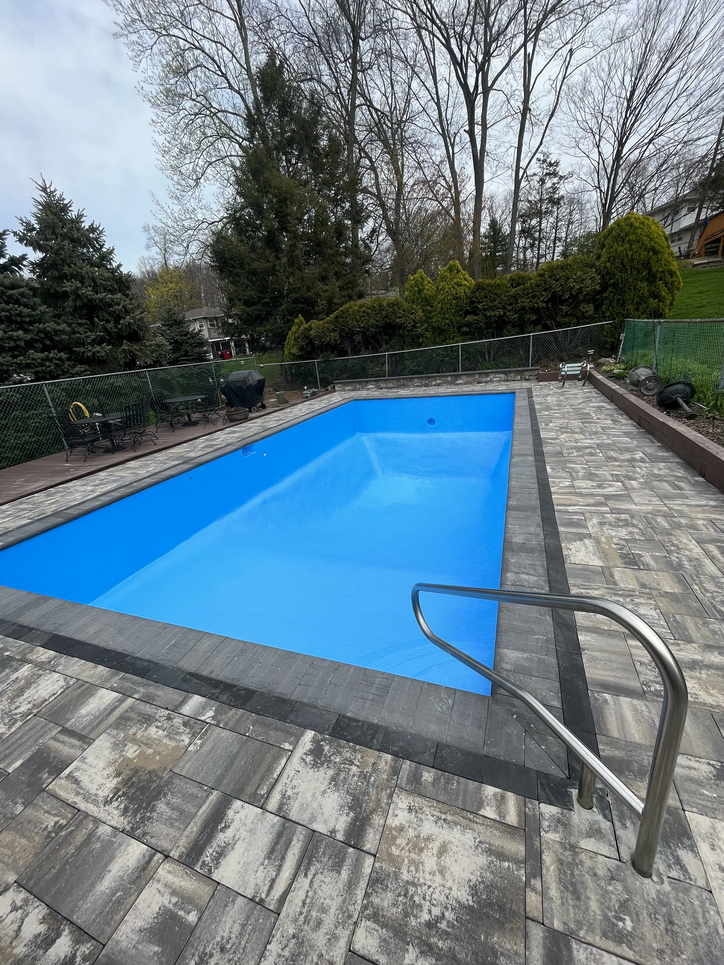 Empty backyard swimming pool with blue interior and metal handrail, surrounded by stone paving and a fence, with trees and bushes in the background.