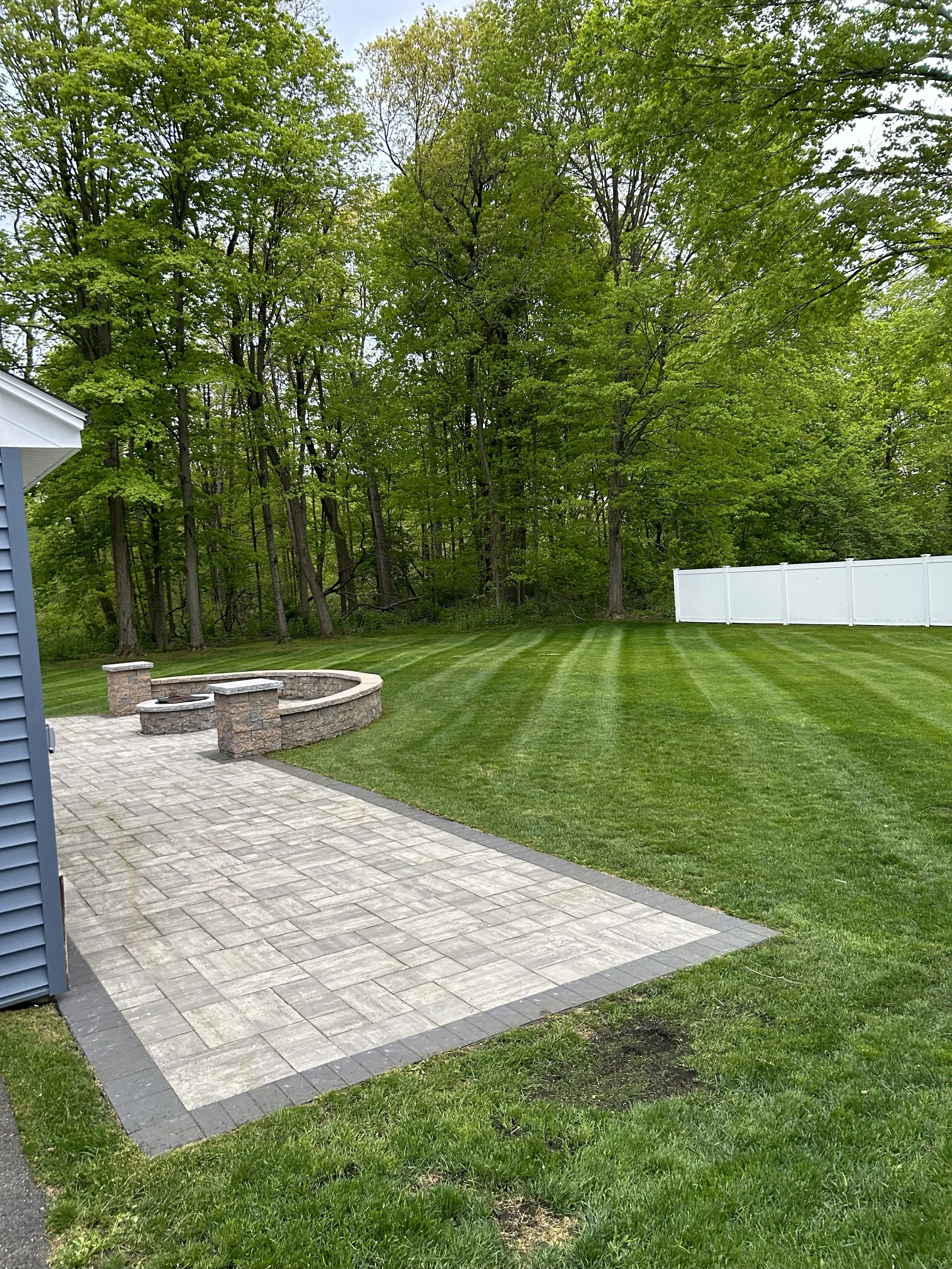 Backyard with a stone patio, fire pit, and neatly mowed lawn surrounded by trees and a white fence.