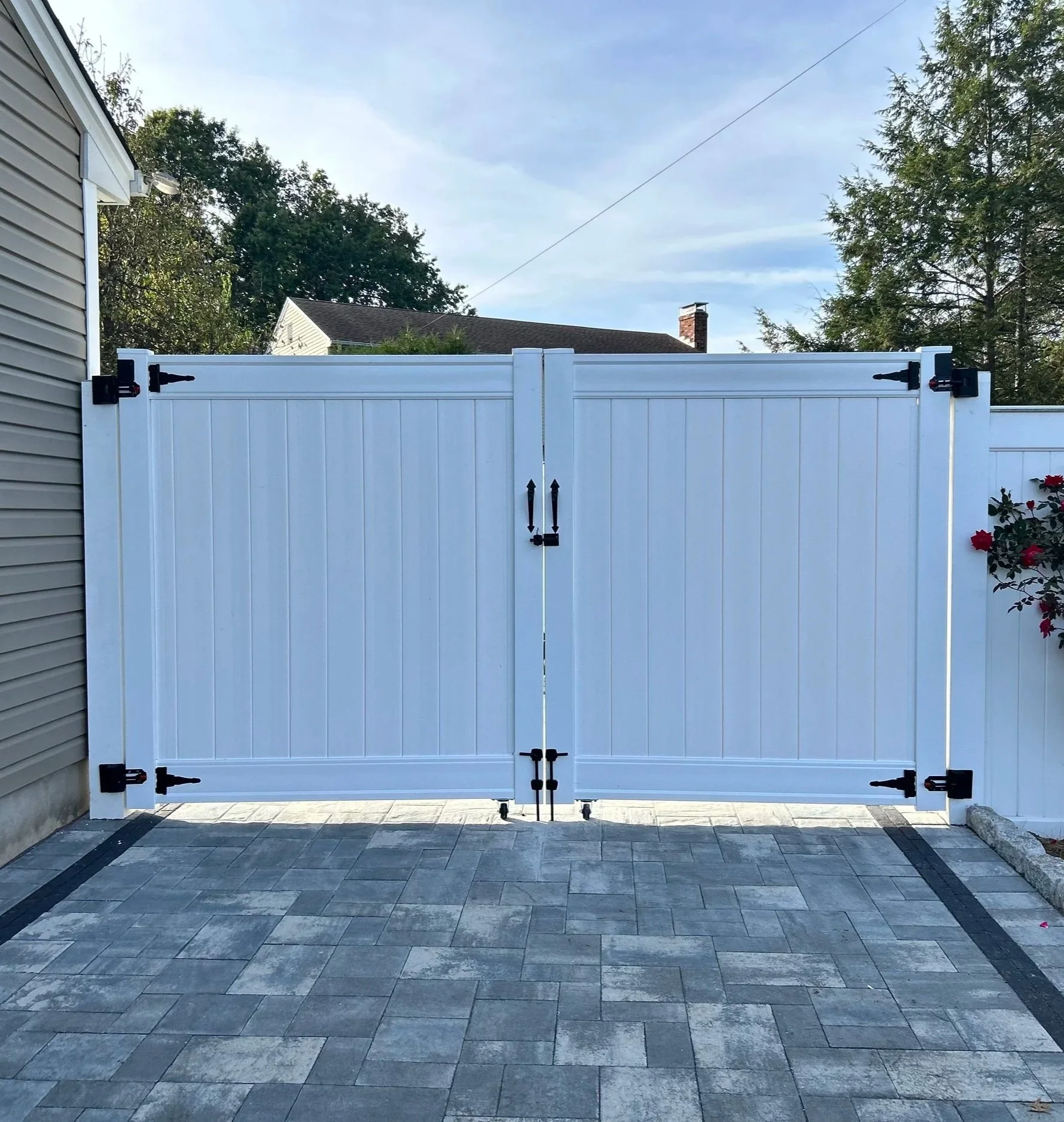 White double gate with black hinges and latch, closed in front of a paved driveway. A house with beige siding is on the left, and a bush with red flowers is on the right. Trees and a partly cloudy sky are in the background.