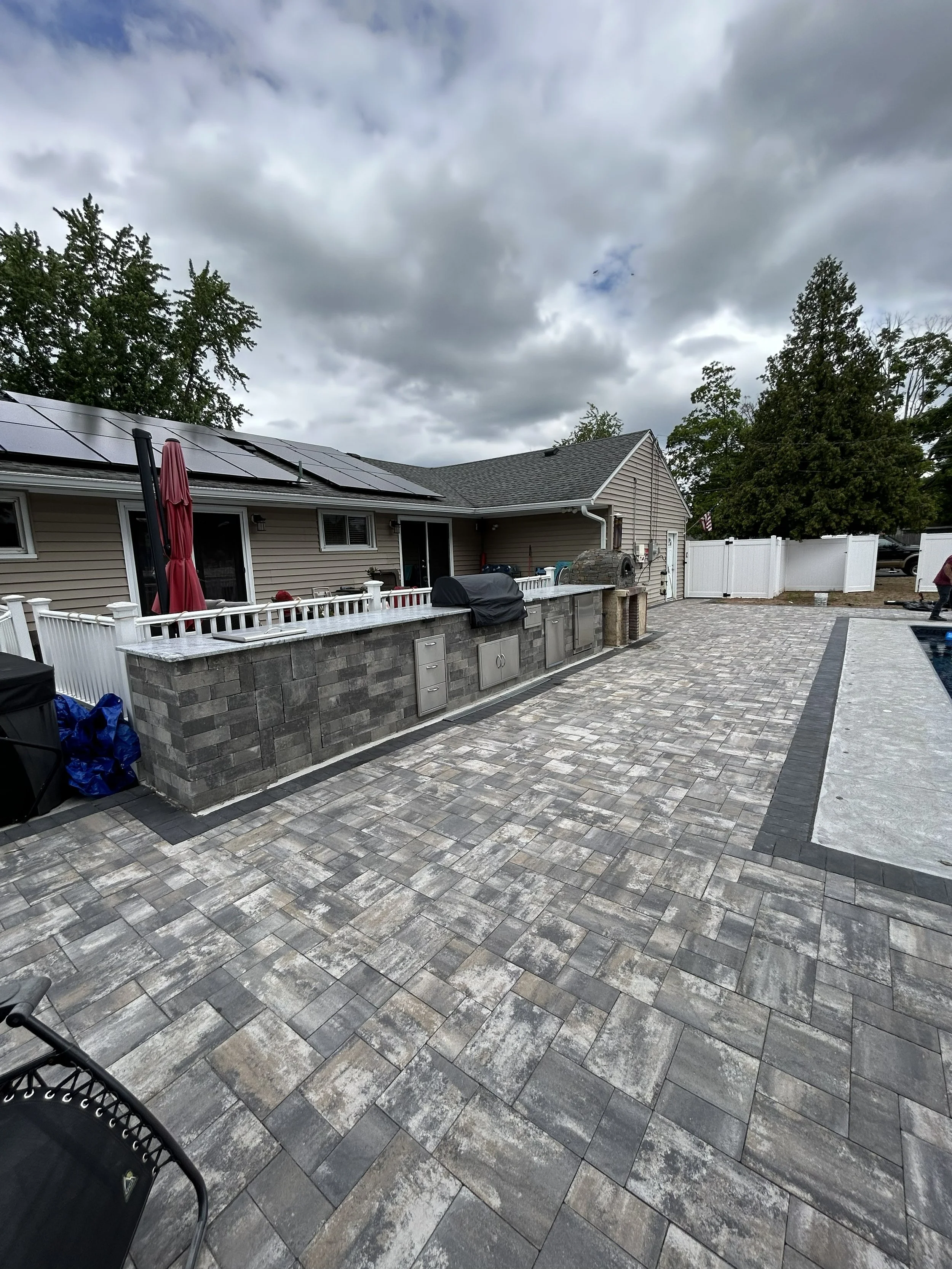 Backyard patio with gray brick pavers, an outdoor kitchen with stainless steel appliances, a grill covered by a black cover, and a white railing. There is a house with tan siding, solar panels on the roof, and a cloudy sky overhead.