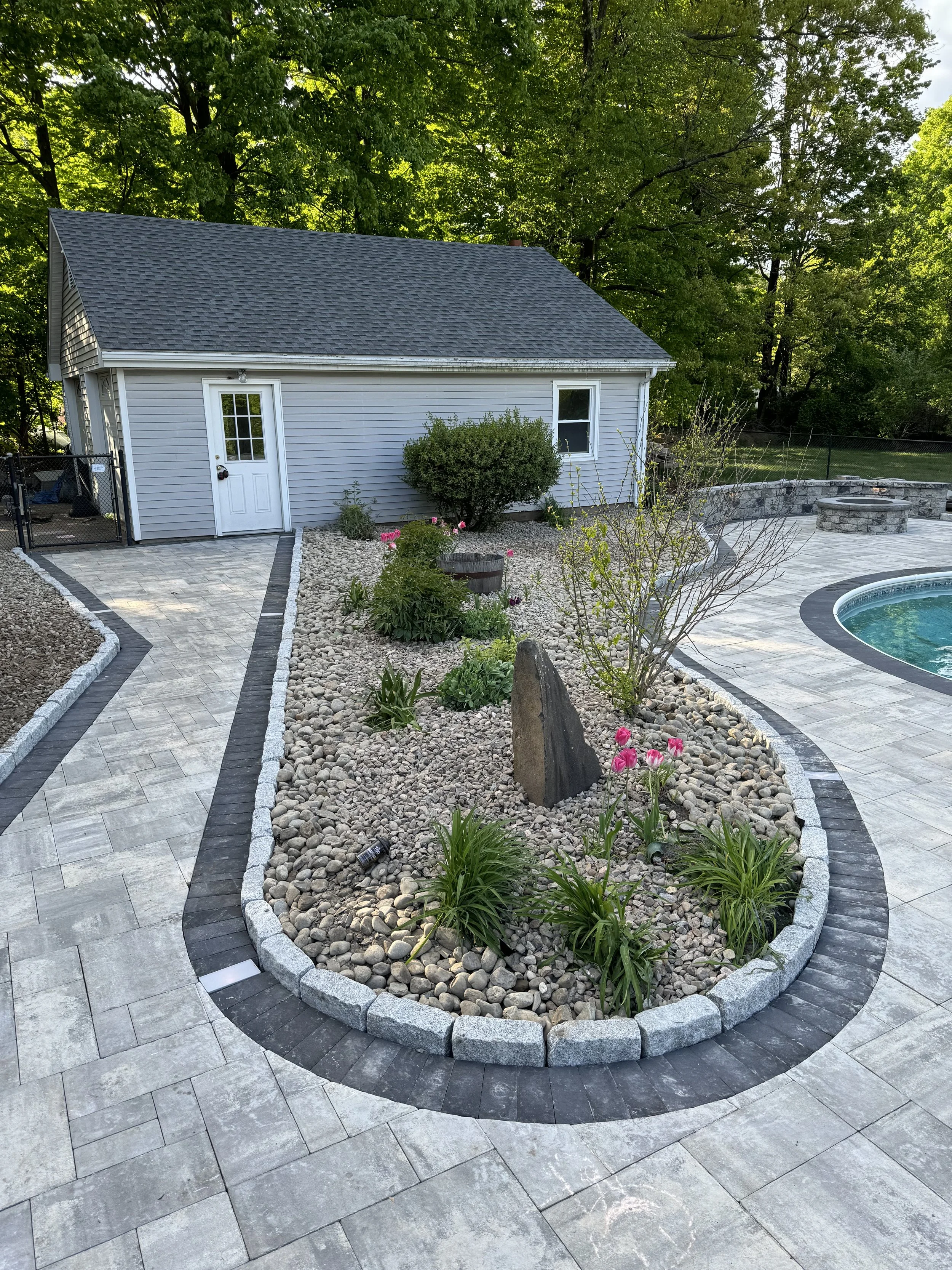 Backyard with a paved patio, in-ground pool, garden bed with stones, plants, flowers, and a small white house with gray siding and a dark gray shingle roof.