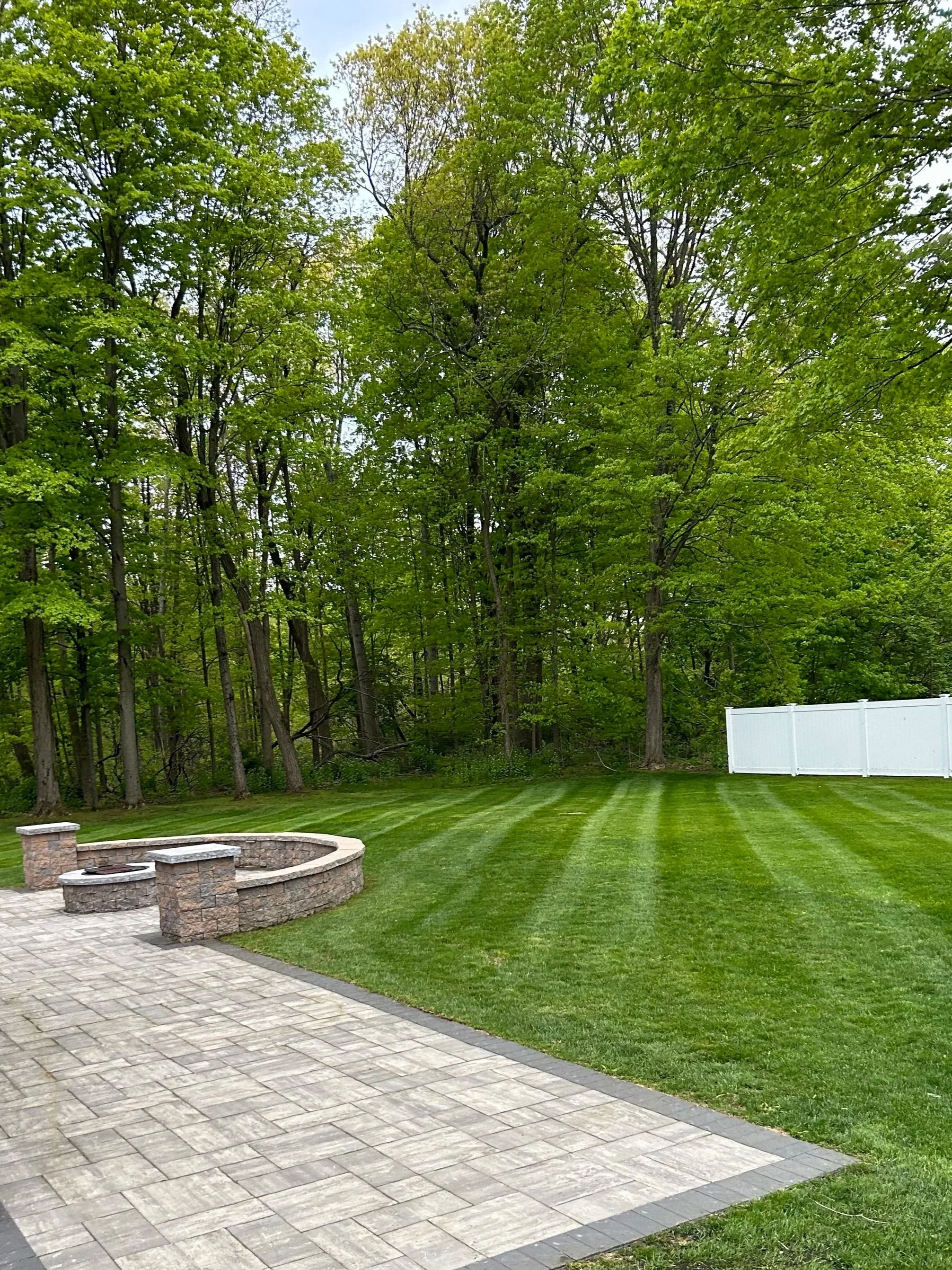 A backyard with a paved patio, a stone fire pit, a well-manicured lawn with striped grass, and tall green trees in the background, enclosed by a white fence.
