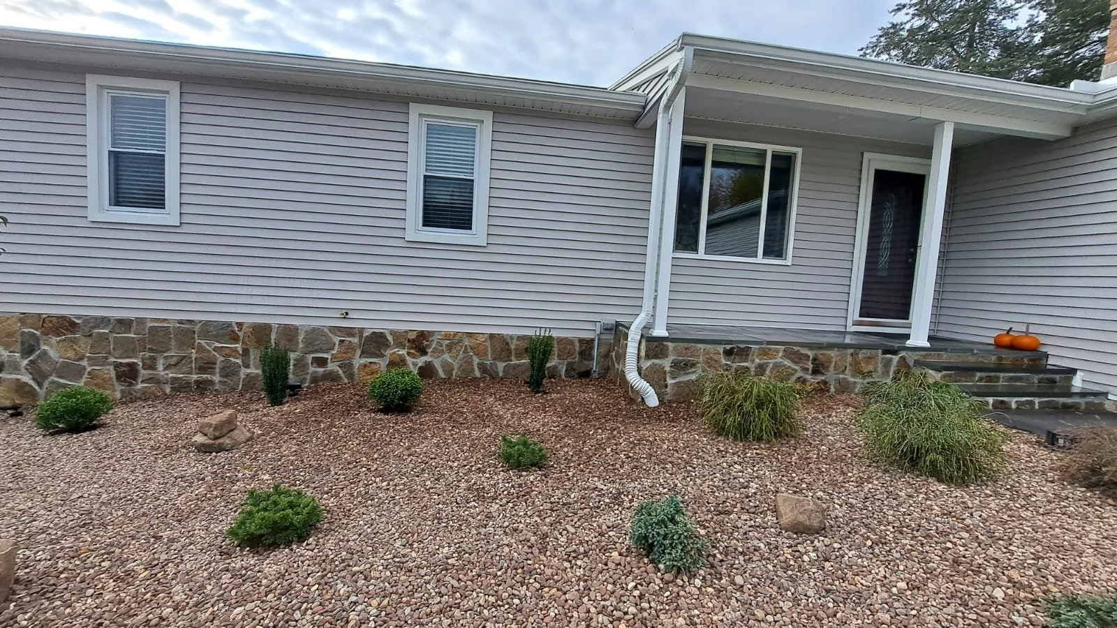 Exterior view of a house with a stone foundation, beige vinyl siding, four windows, a small porch with steps, and pumpkins on the porch steps surrounded by small bushes and gravel landscape.