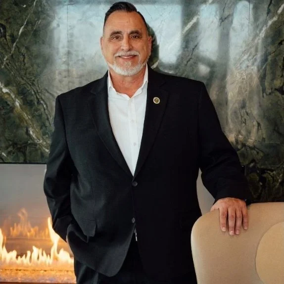 A man in a black suit standing indoors with a marble wall background and a beige chair beside him.