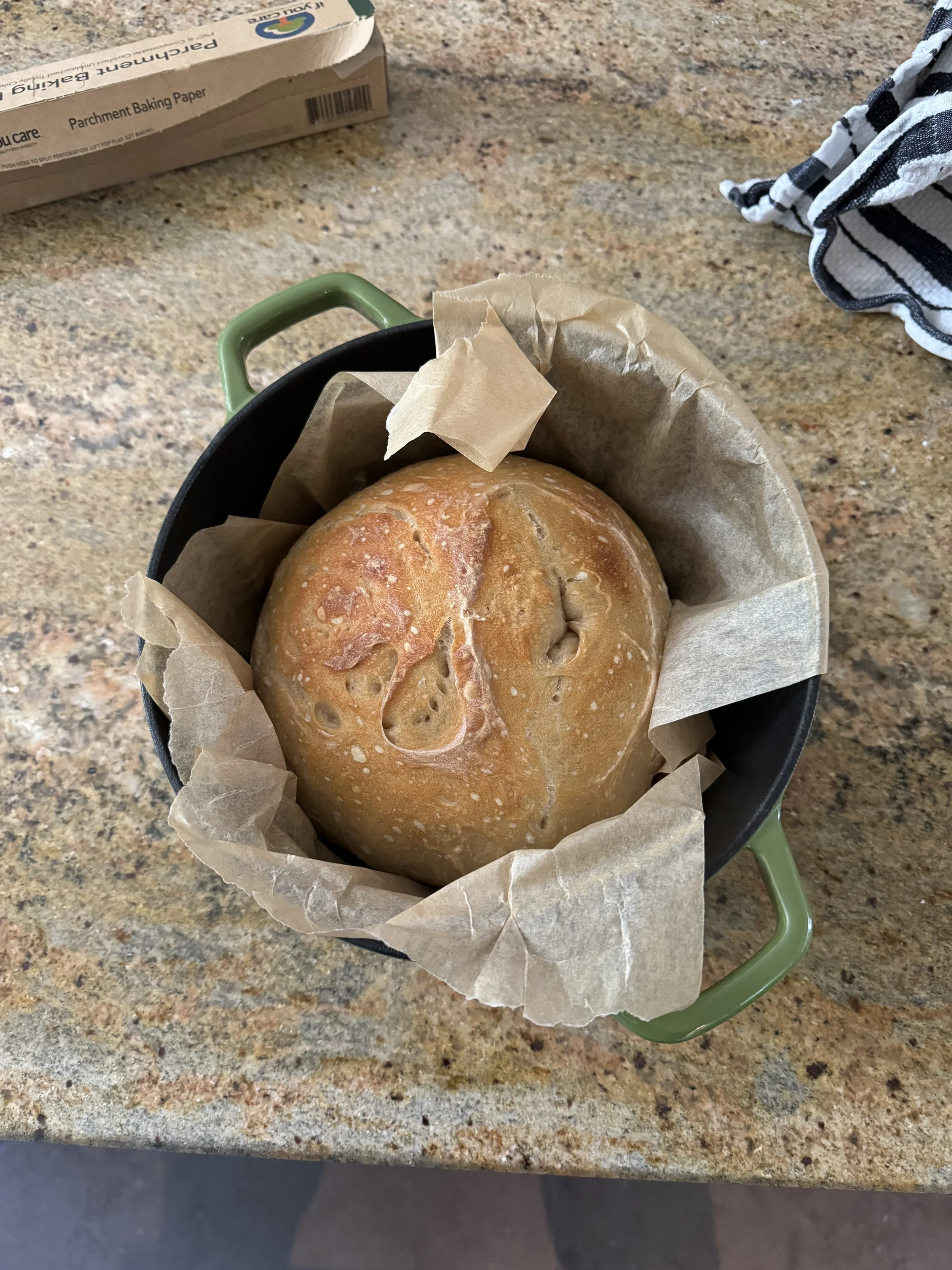 A round bread roll in a small black pan lined with parchment paper on a speckled kitchen countertop.