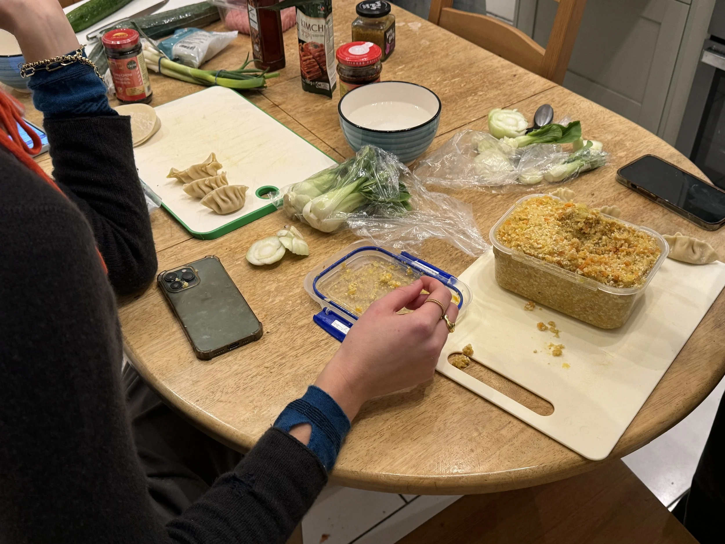 Person preparing dumplings on a kitchen table with ingredients including green onions, garlic, and various jars and containers of spices and seasonings.