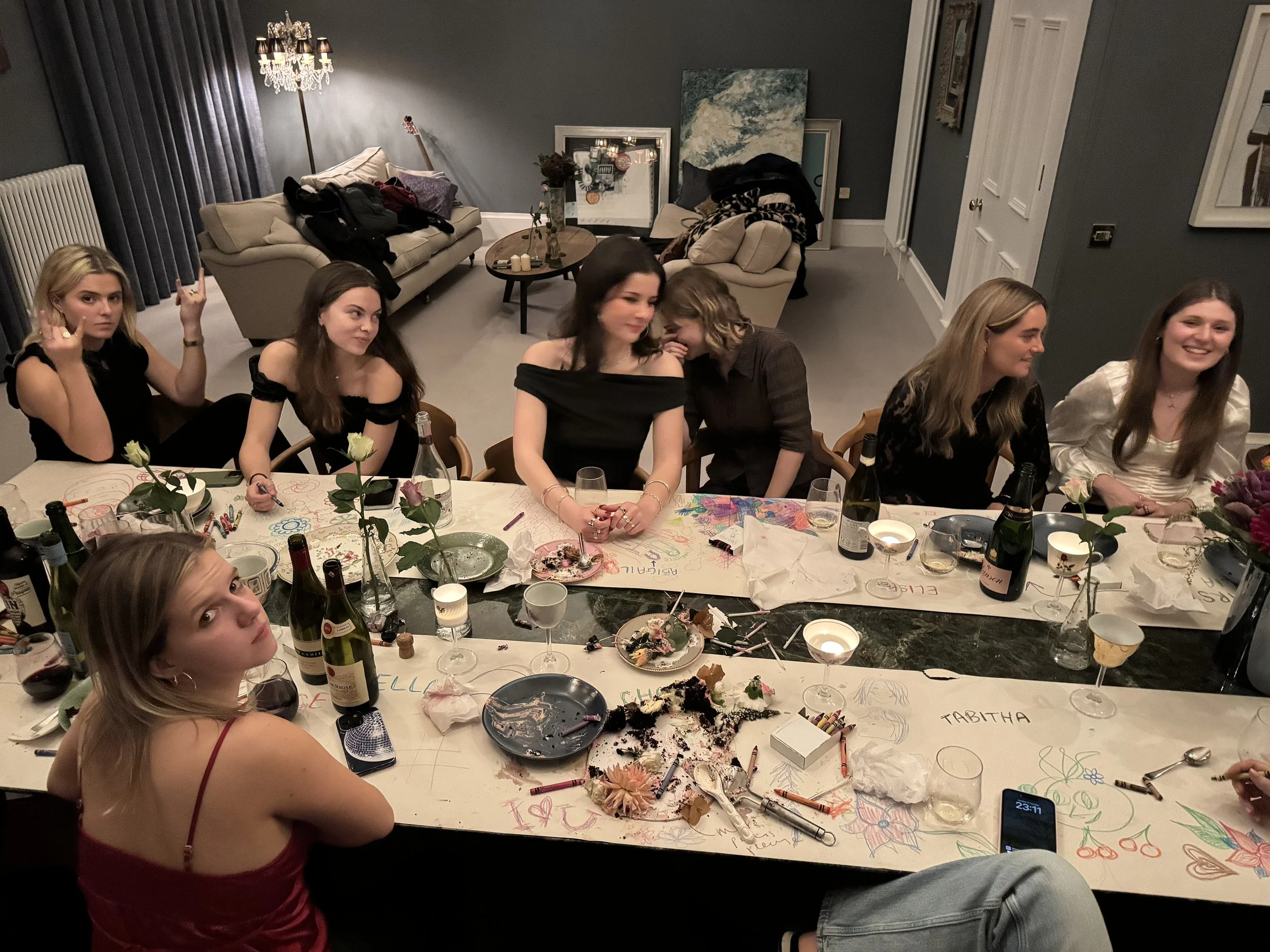 A group of young women sitting and standing around a decorated table after a celebration, with leftover food, drinks, and drawings on the table and wall.