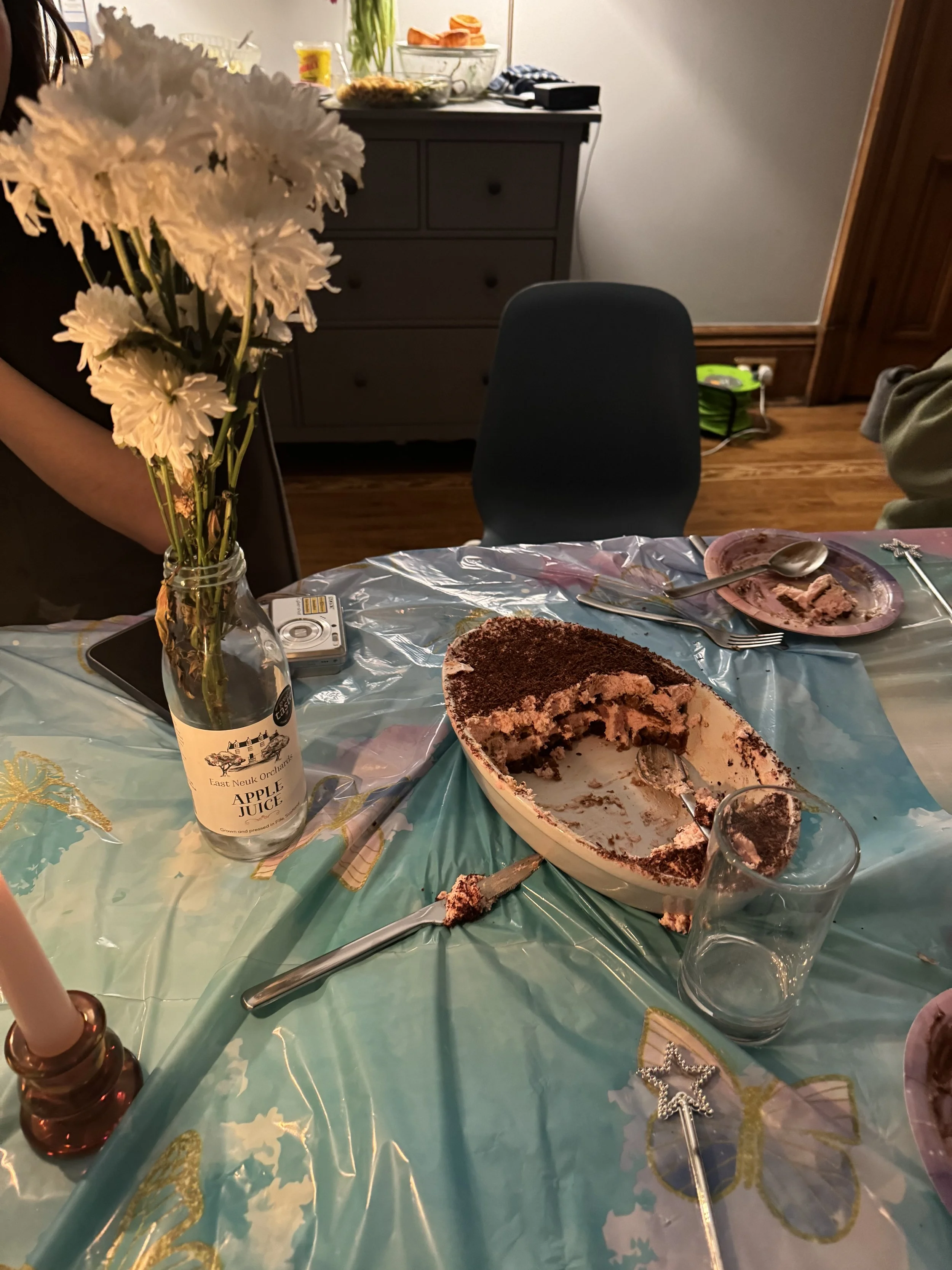 A partially eaten chocolate and vanilla layered cake on a blue tablecloth, with a glass, cake-serving utensil, and decorative star wand nearby. A vase with white flowers, a bottle labeled 'Apple Juice,' and a picture camera are also on the table. In the background, kitchen items and a black chair are visible.