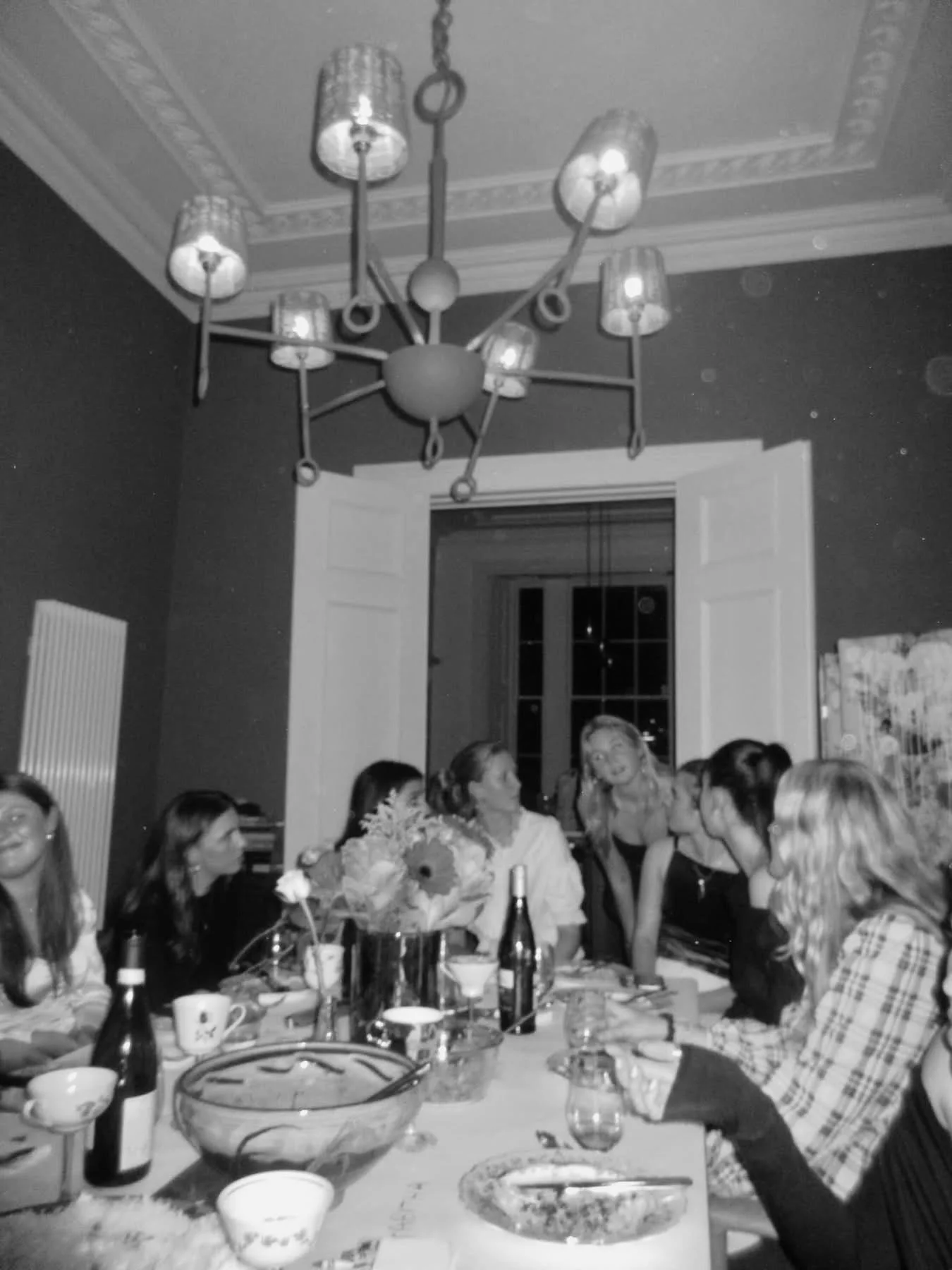 A black and white photo of a group of people gathered around a dining table in a decorated room, with a chandelier hanging from the ceiling.