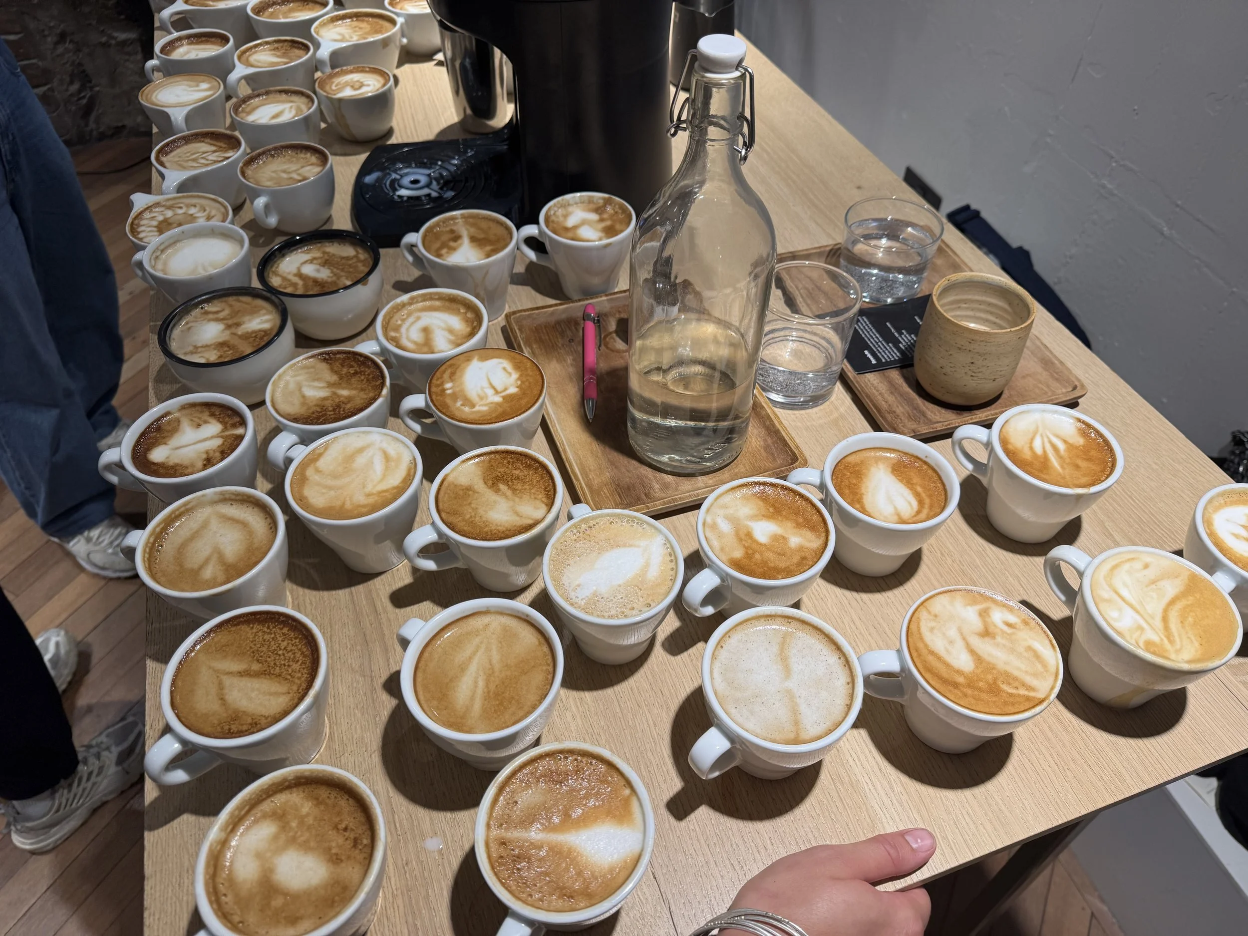 A wooden table filled with numerous cups of coffee with latte art, a glass bottle, several glasses of water, and a pink pen.