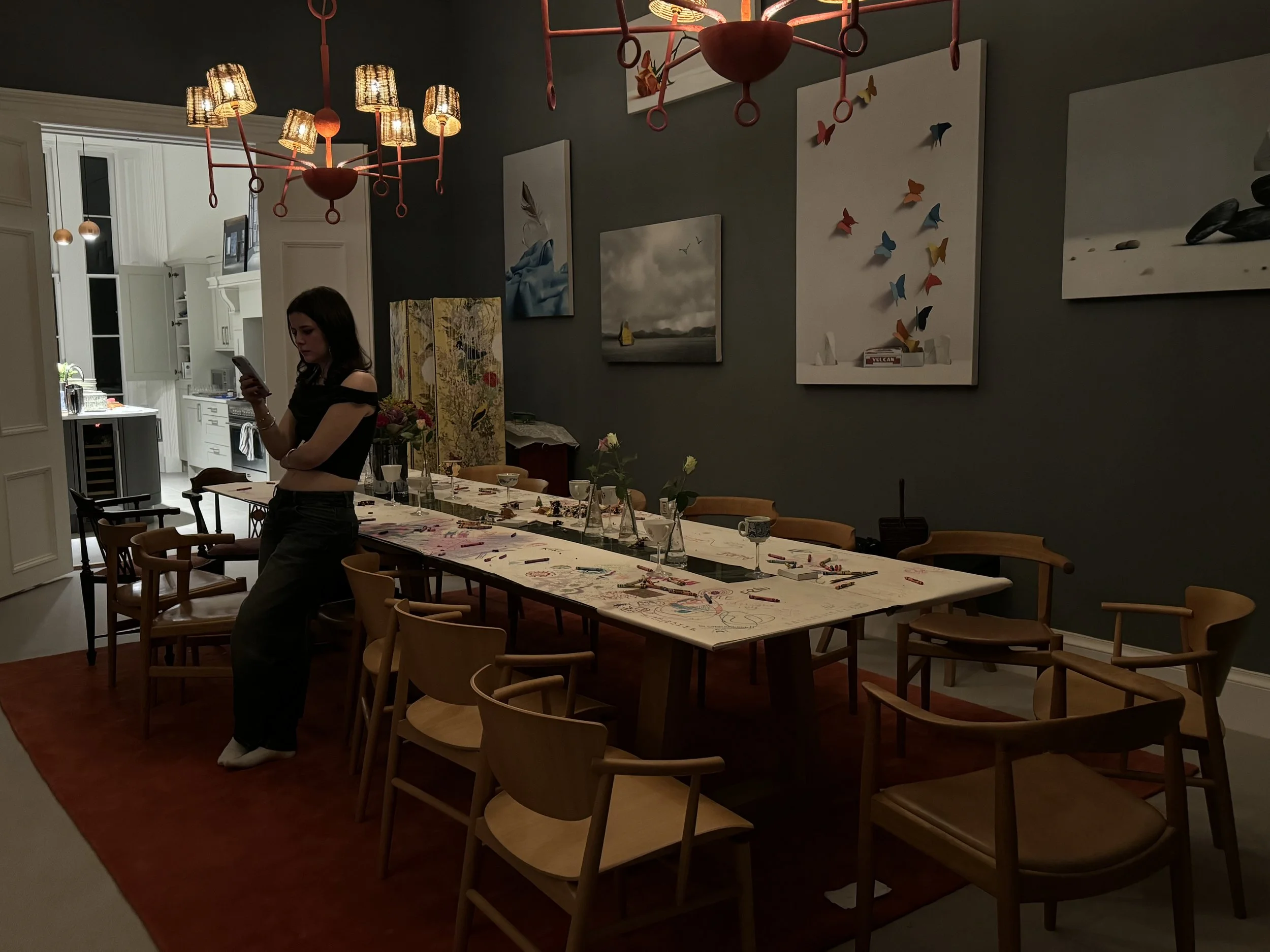 A woman in black clothing standing by a long dining table in a dimly lit dining room, looking at her phone. The table has art supplies and decorations, with chairs arranged around it. The room features hanging lamps, artwork on the walls, and an open kitchen space in the background.