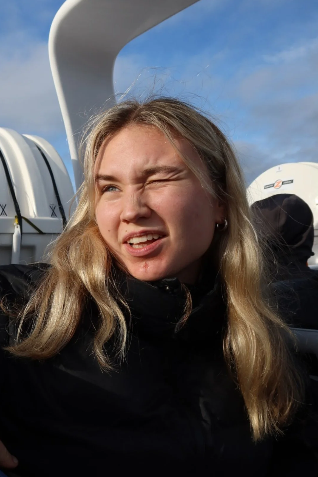 A woman with blonde hair making a squinting face, outdoors with a blue sky and white clouds behind her.