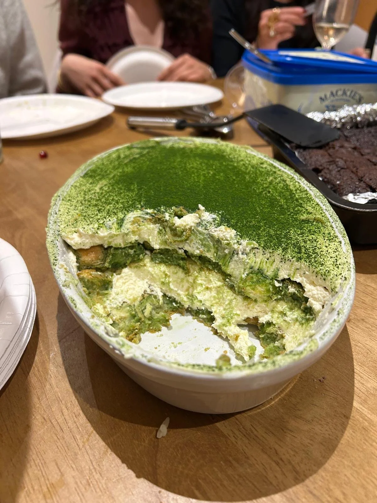 A partially eaten green tea tiramisu in a white bowl on a wooden table surrounded by plates, a blue-lidded dessert container, and blurred figures of people dining.