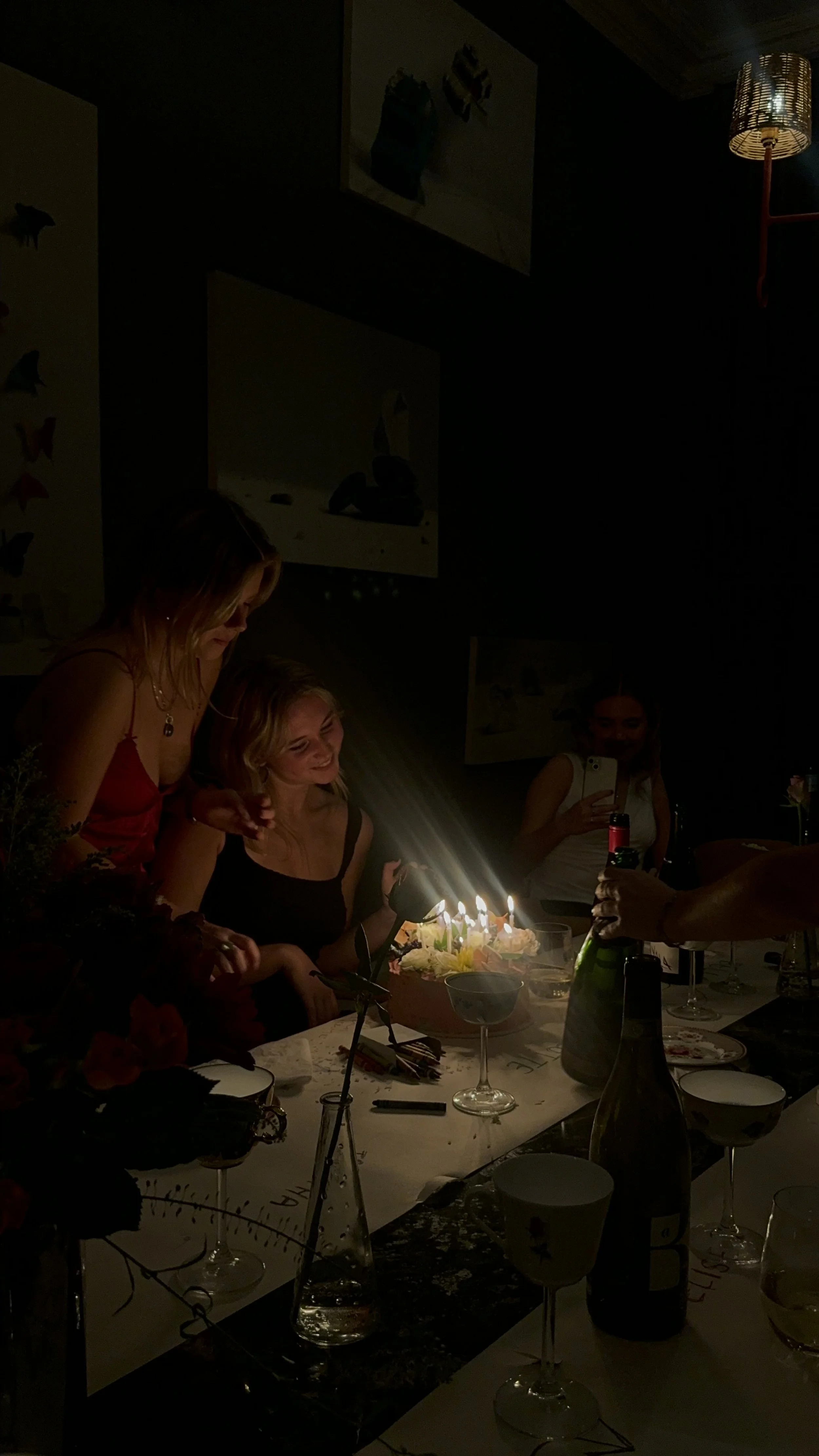 Group of women celebrating a birthday with a cake, lit candles, and drinks in a dimly lit room.