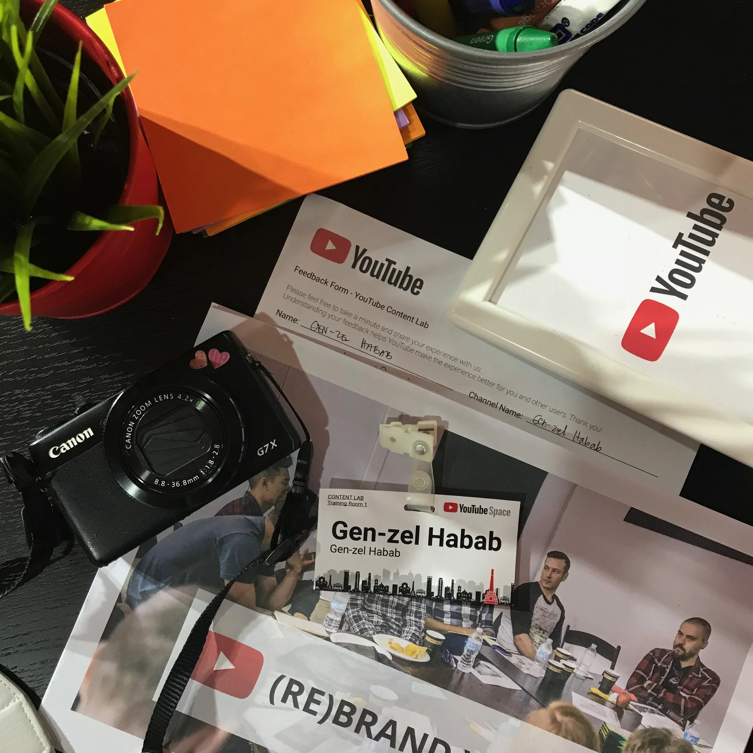 A table with YouTube branding materials, a Canon camera, a plant in a red pot, colorful sticky notes, a metal container with markers, a YouTube Feedback Form, a YouTube Space badge with the name Gen-zel Habab, and a photo of a group of people in a me