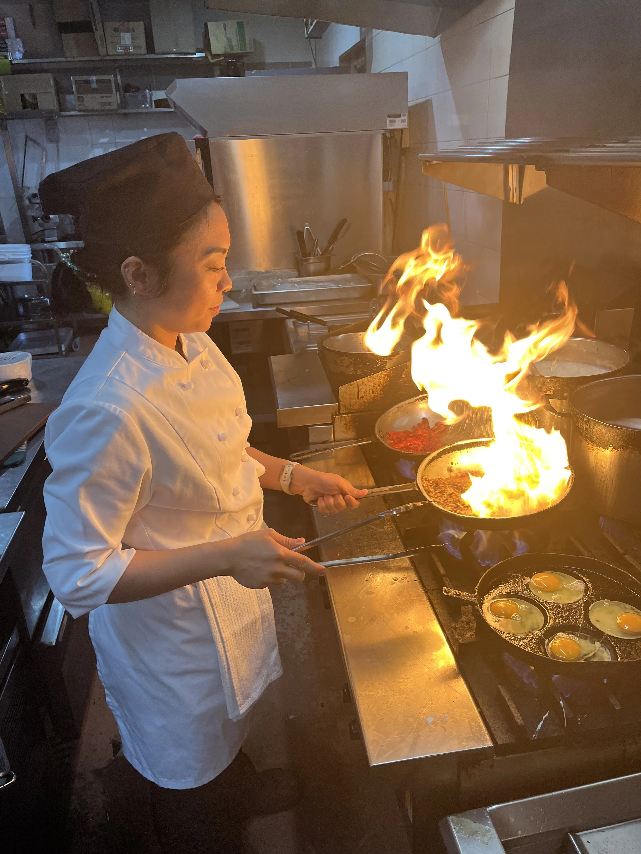 A female chef in a white uniform and black chef's hat cooking eggs and other dishes on a stove, with flames rising from a pan.