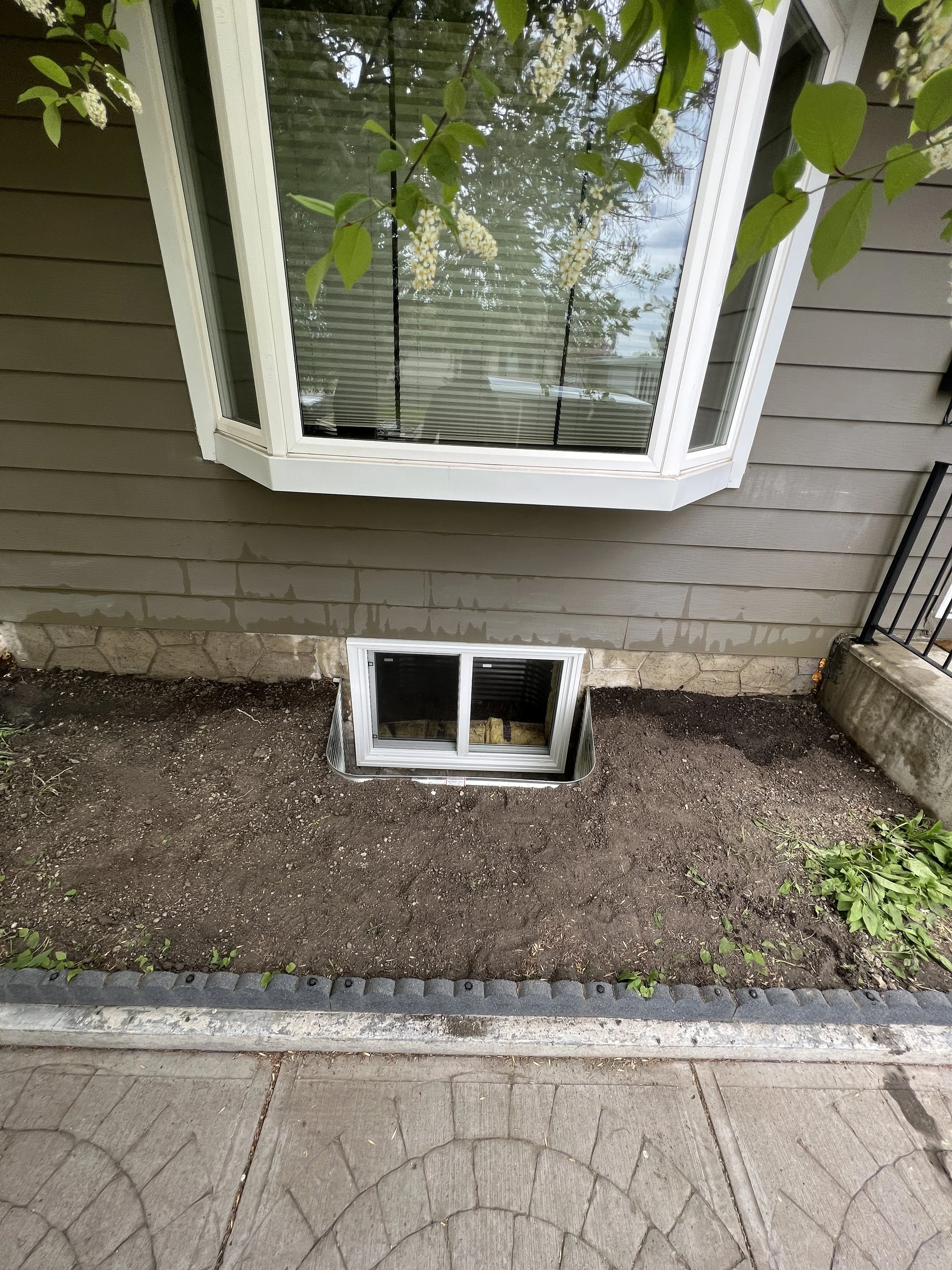 A large basement window installed below a larger bay window on the exterior of a house. The ground around the basement window is freshly excavated with no grass or landscaping yet.