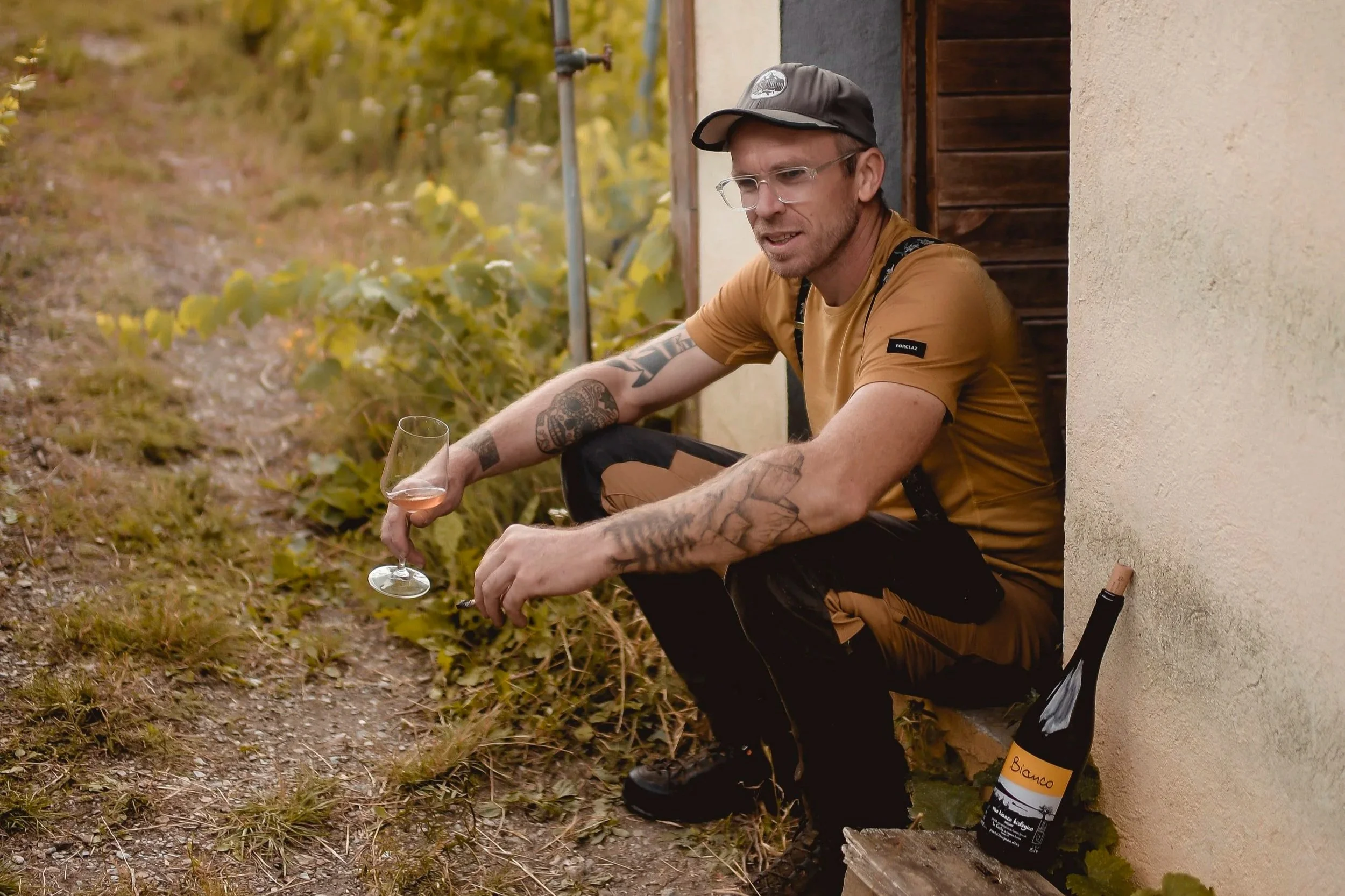 Julien Guillon with tattoos on his arms, wearing glasses, a baseball cap, and a mustard-colored T-shirt, sits outside near a wall holding a glass of wine. There is a wine bottle with a yellow label on the ground beside him.
