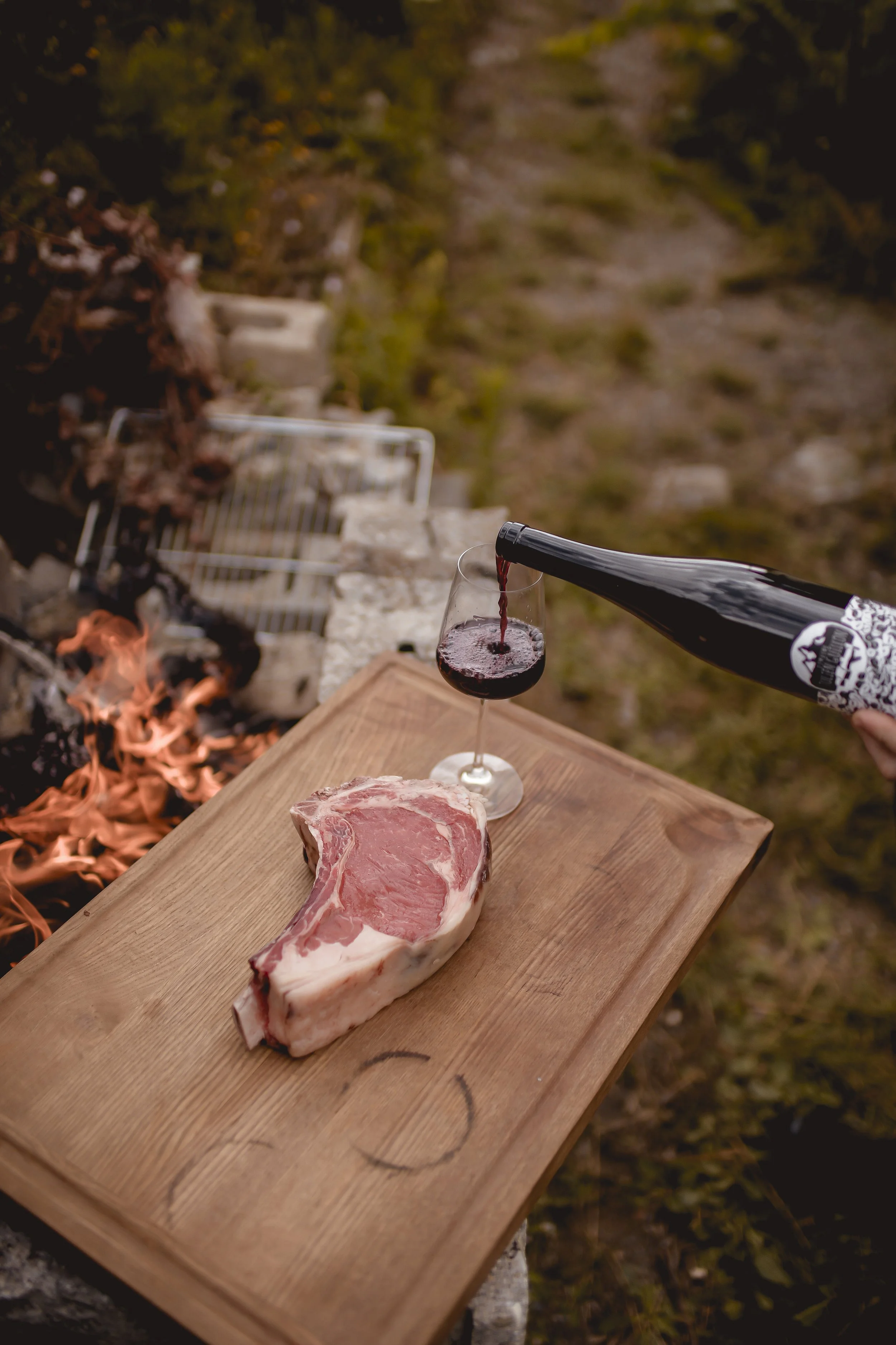 A piece of raw steak on a wooden cutting board with a glass of red wine being poured from a bottle outdoors, with a fire and grill in the background.