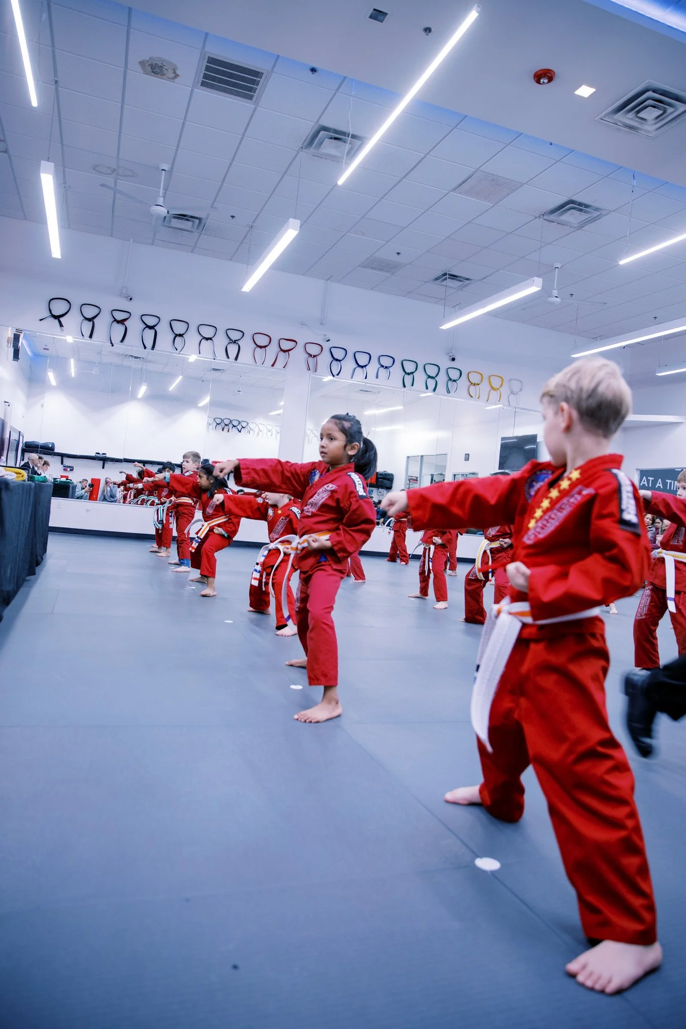 Children practicing martial arts in a dojo, all wearing red uniforms and white belts, performing a synchronized kick in front of mirrors and colorful training equipment.