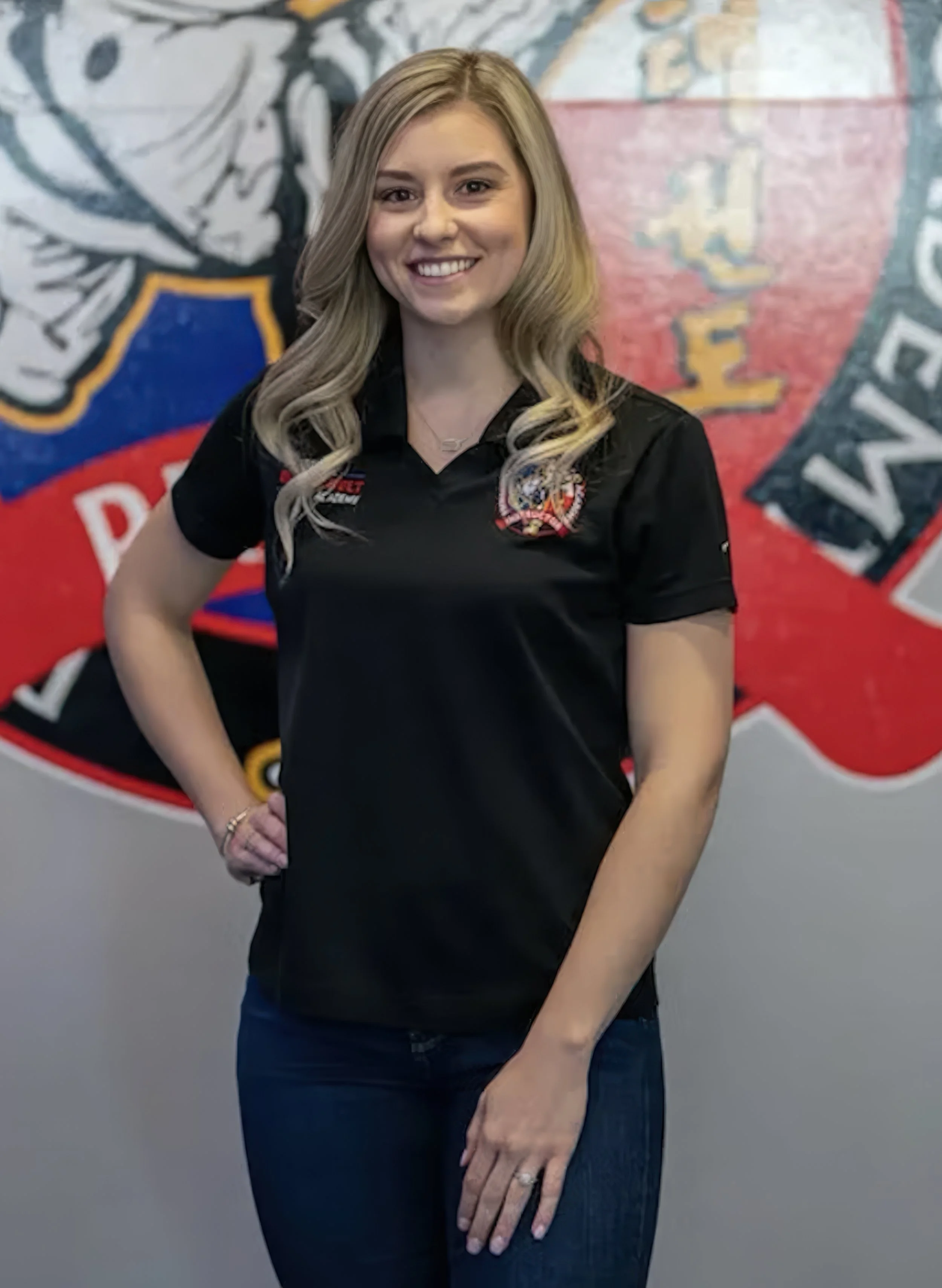 A young woman with long, wavy blonde hair smiles and poses with her hand on her hip in front of a colorful mural.