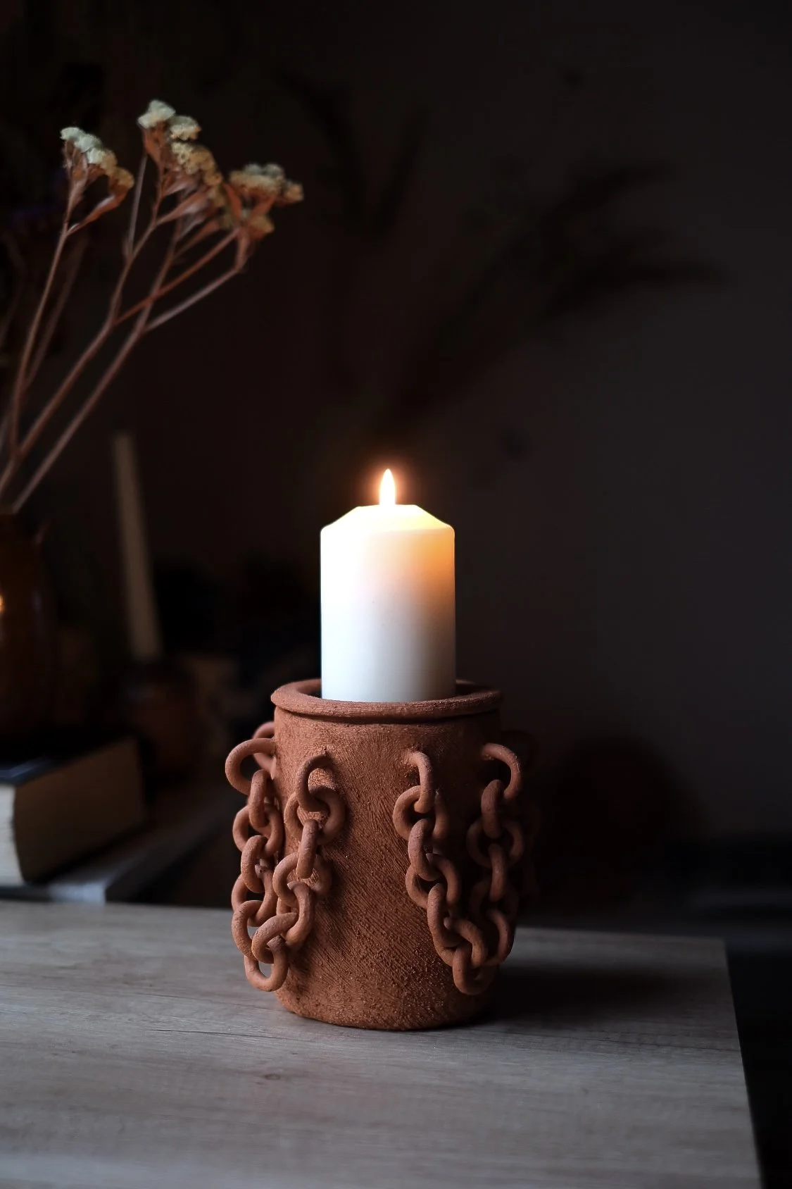 A white candle burning in a terracotta holder with chain links, placed on a wooden surface with dried flowers in a vase in the background.