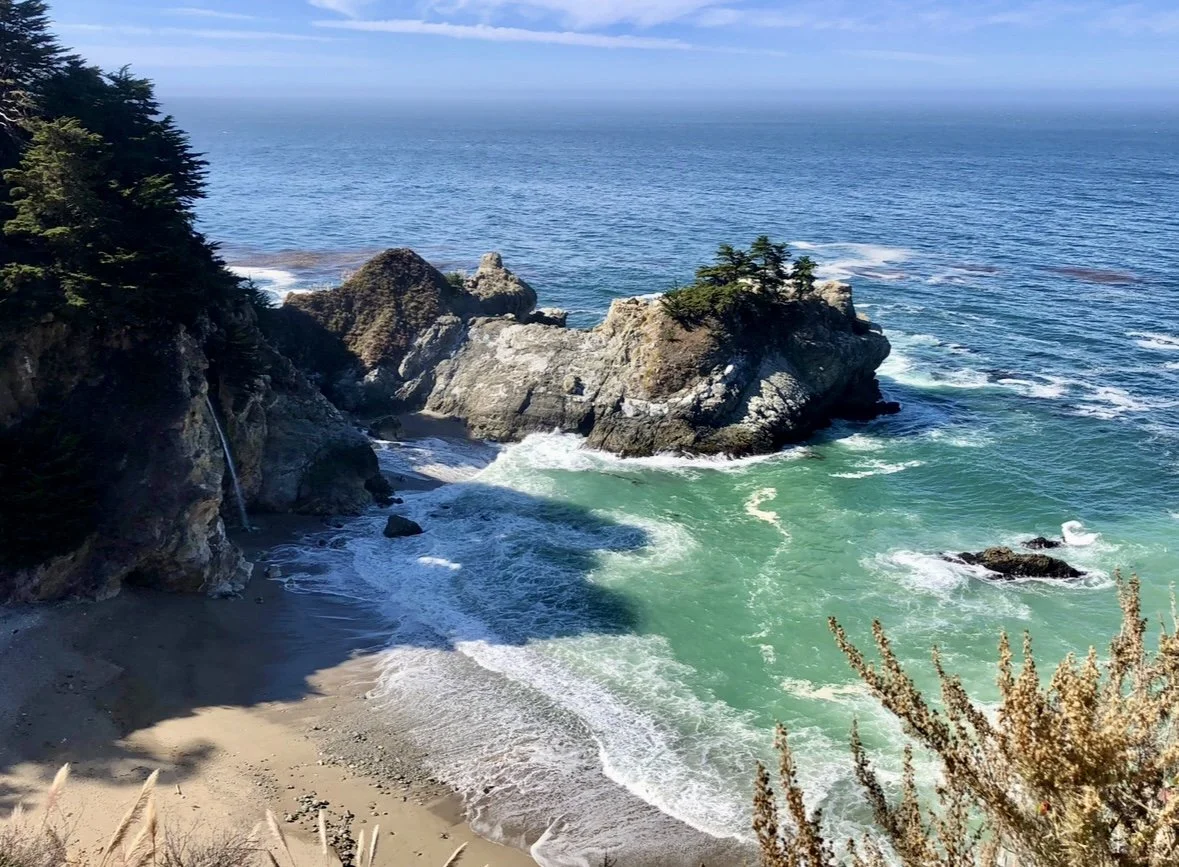 Scenic view of a rocky coastline of Big Sur with trees, waves crashing on the shore, and the ocean extending to the horizon under a blue sky.