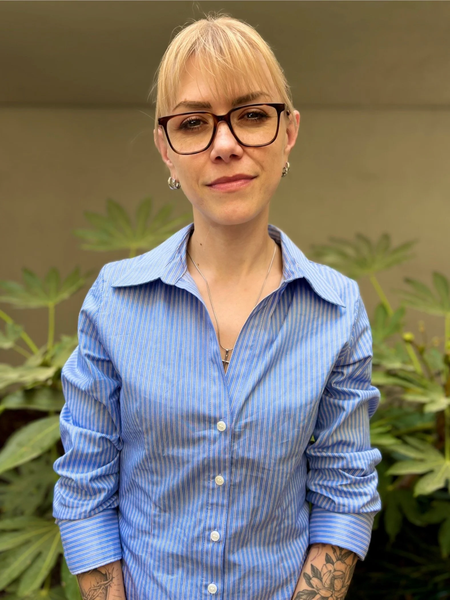 A woman with blonde hair, wearing glasses, a blue striped button-up shirt, and jewelry, standing in front of green plants.
