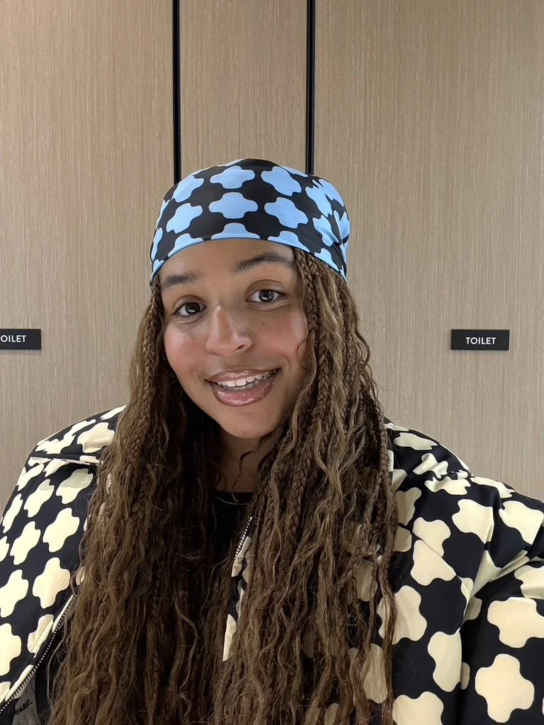Woman with long curly hair wearing a black and white patterned jacket and a black and blue patterned headscarf, standing in front of an elevator with signs indicating toilets.