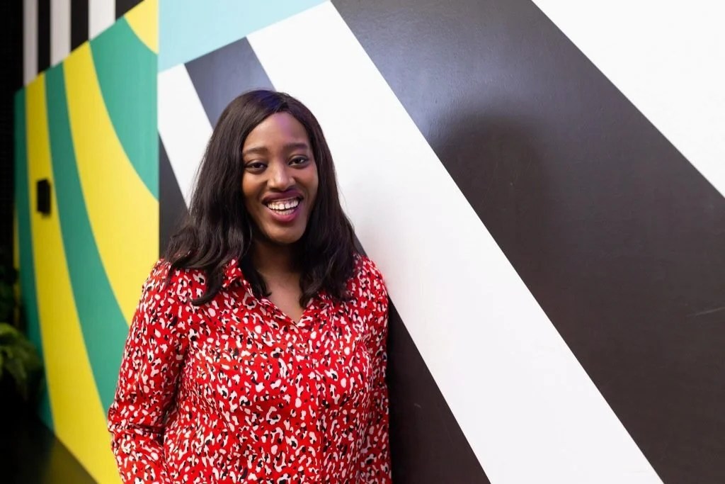 A woman with long dark hair smiling, wearing a red and white patterned blouse standing next to a black and white geometric mural.