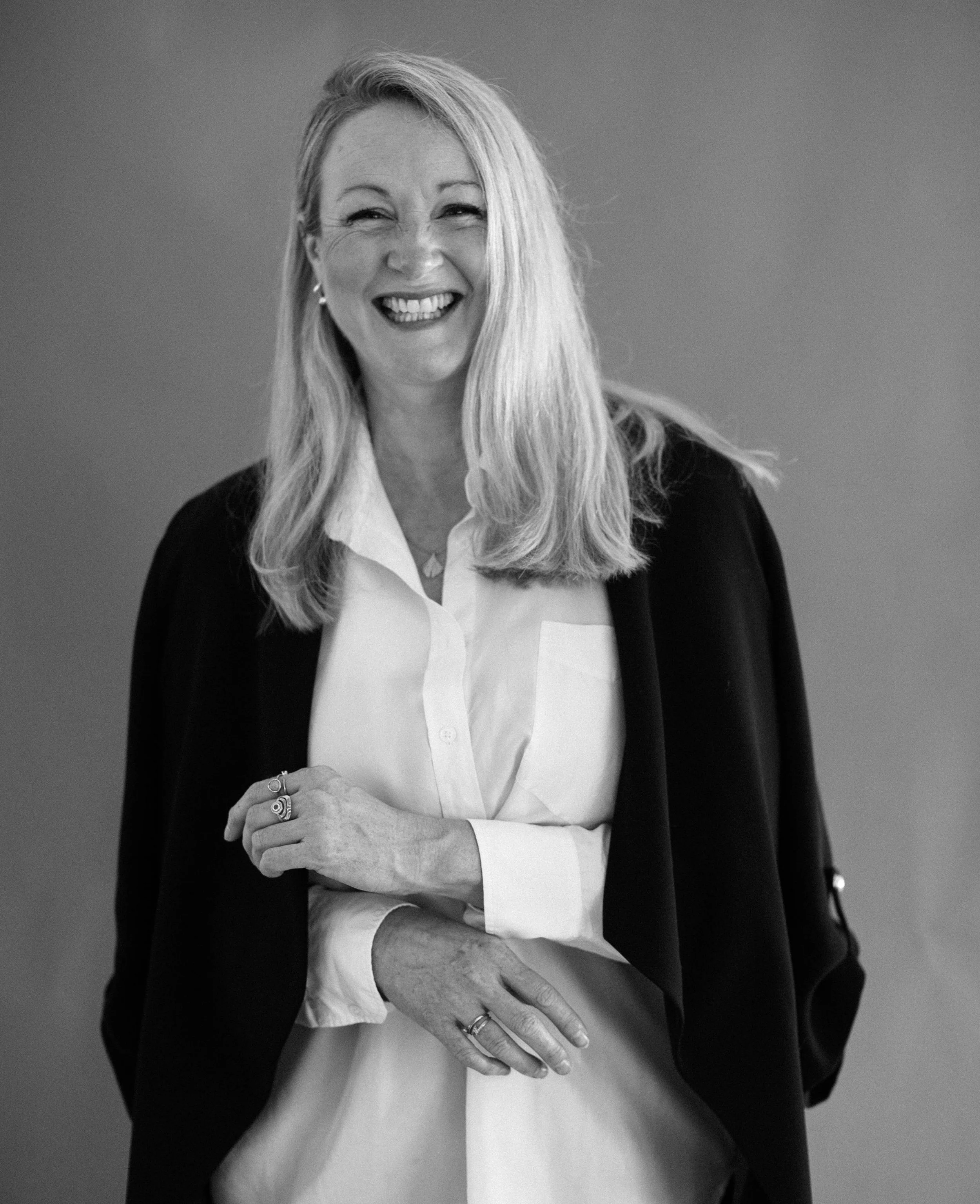 Black and white portrait of a smiling woman with long blonde hair, wearing a white blouse and a dark blazer, standing against a plain background.