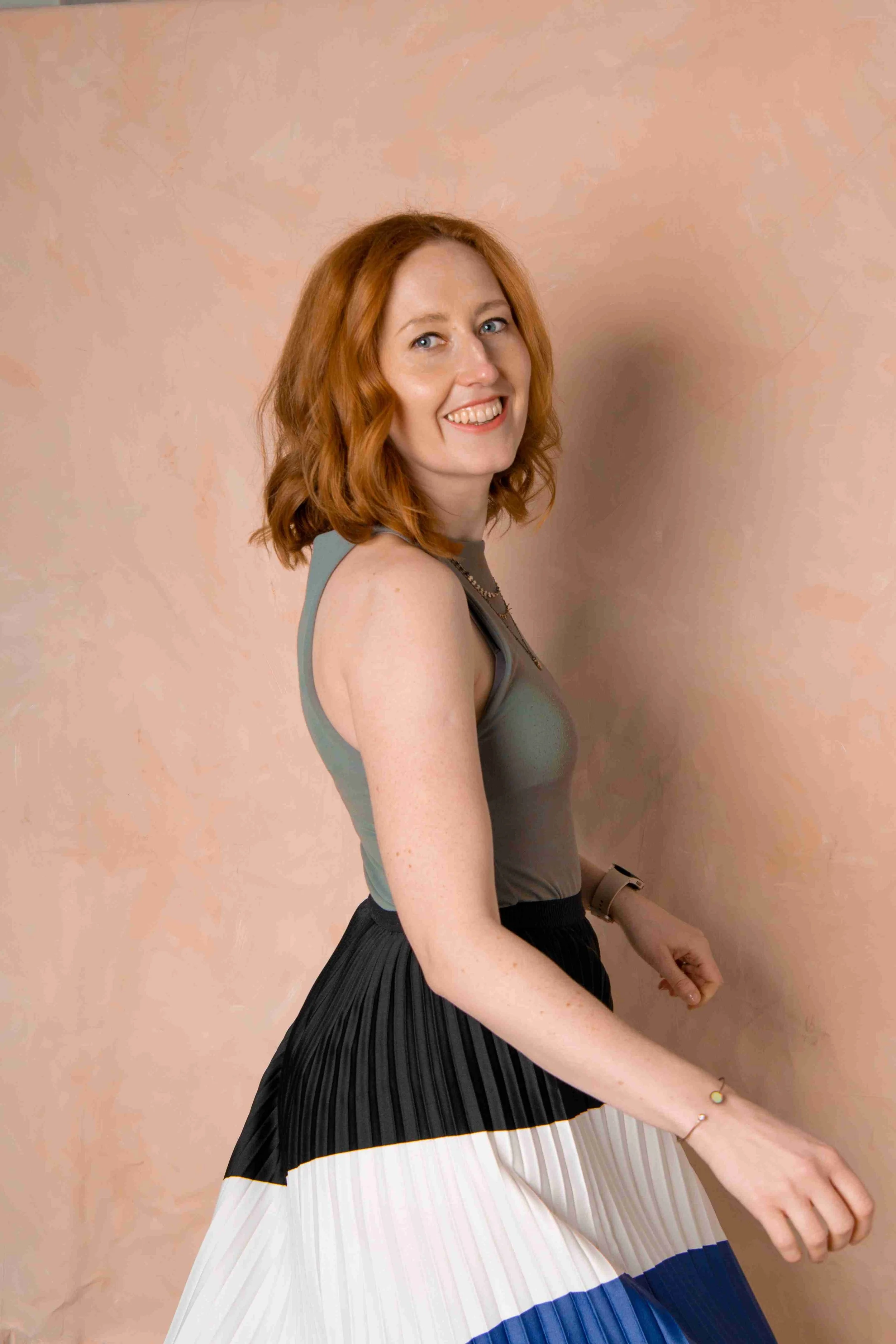 A woman with red hair smiling, wearing a sleeveless gray top and a color-blocked pleated skirt, standing against a pink textured wall.