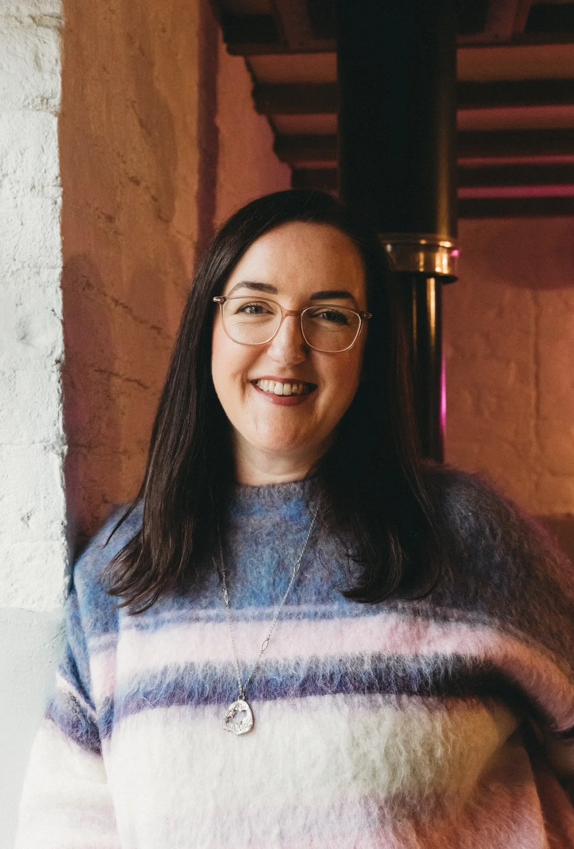 A woman with long dark hair, glasses, and a sweater with pastel stripes, smiling and standing in a warmly lit indoor setting with a brick wall and metal pipe behind her.