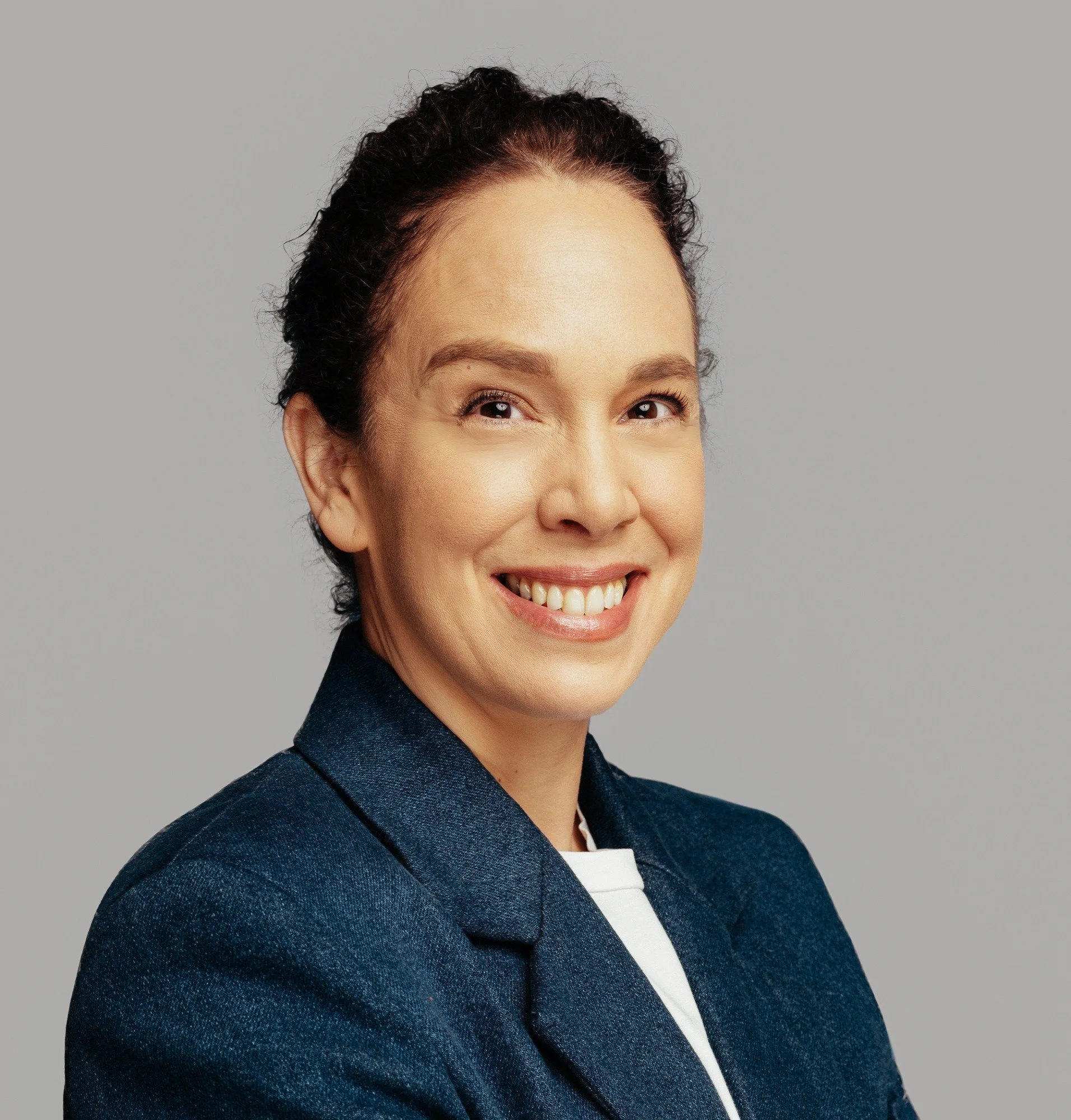 Headshot of a smiling woman with dark hair pulled back, wearing a dark blazer over a white top, standing against a gray background.