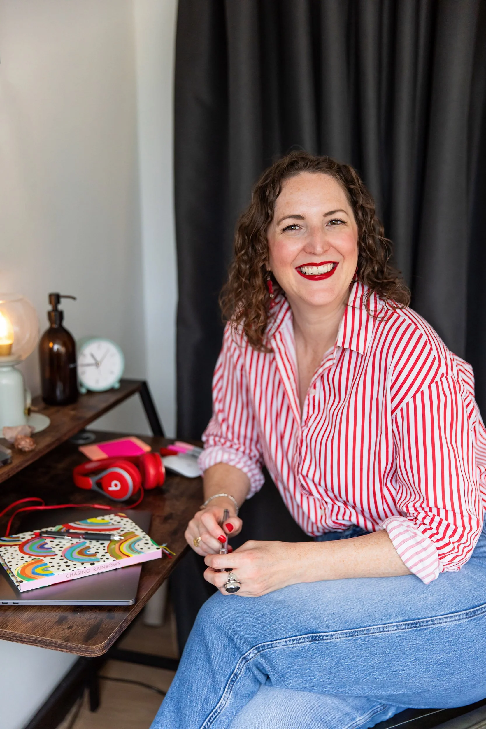 A woman with curly brown hair, wearing a red and white striped shirt and blue jeans, sitting at a desk with a colorful notebook, pink sticky notes, and red headphones. She is smiling and looking at the camera.