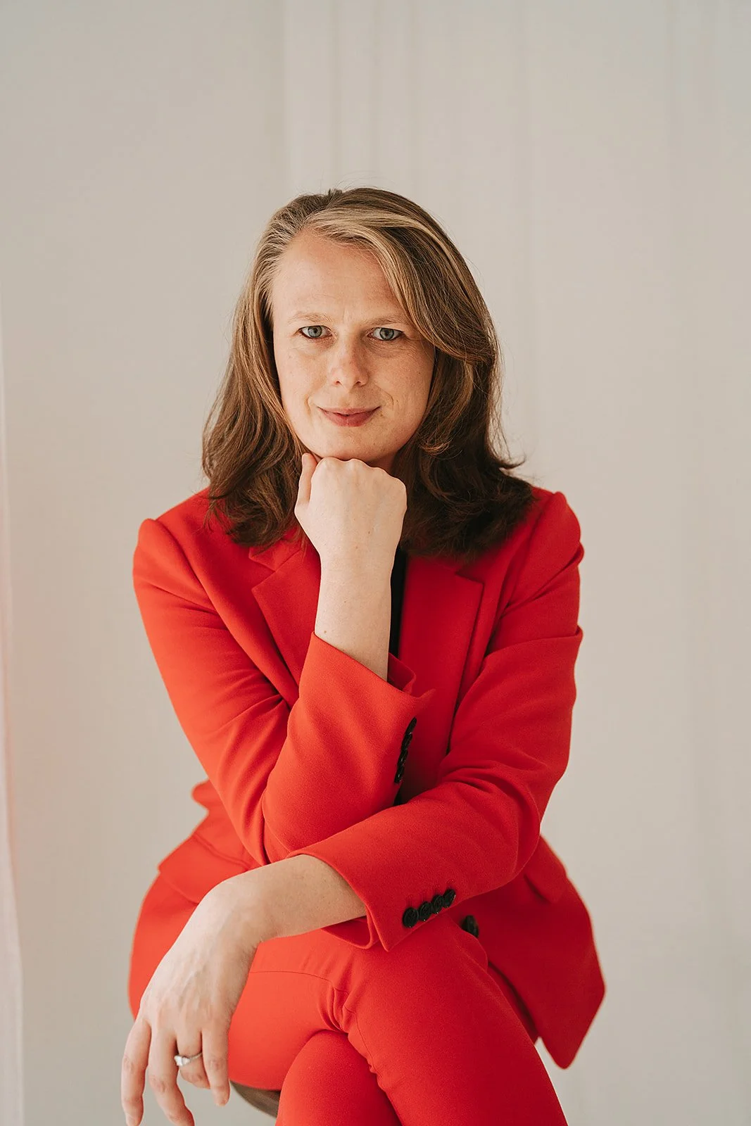 A woman with shoulder-length brown hair and blue eyes wearing a red suit, sitting with her chin resting on her hand, looking thoughtfully at the camera.