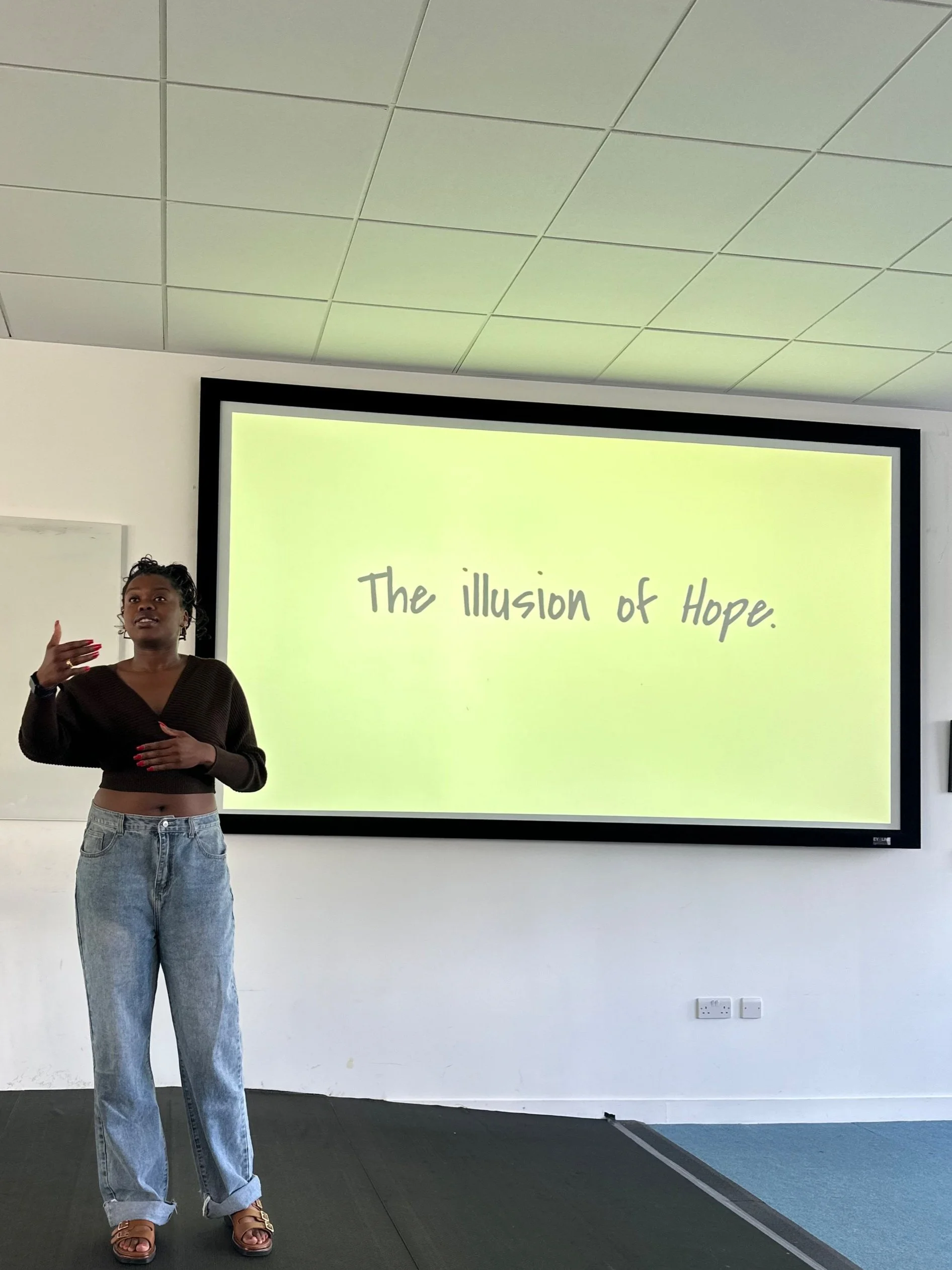 A woman giving a presentation in front of a large screen that displays the words 'The illusion of Hope.' She is wearing a black crop top and high-waisted jeans, standing on a stage in a room with a white wall and ceiling tiles.
