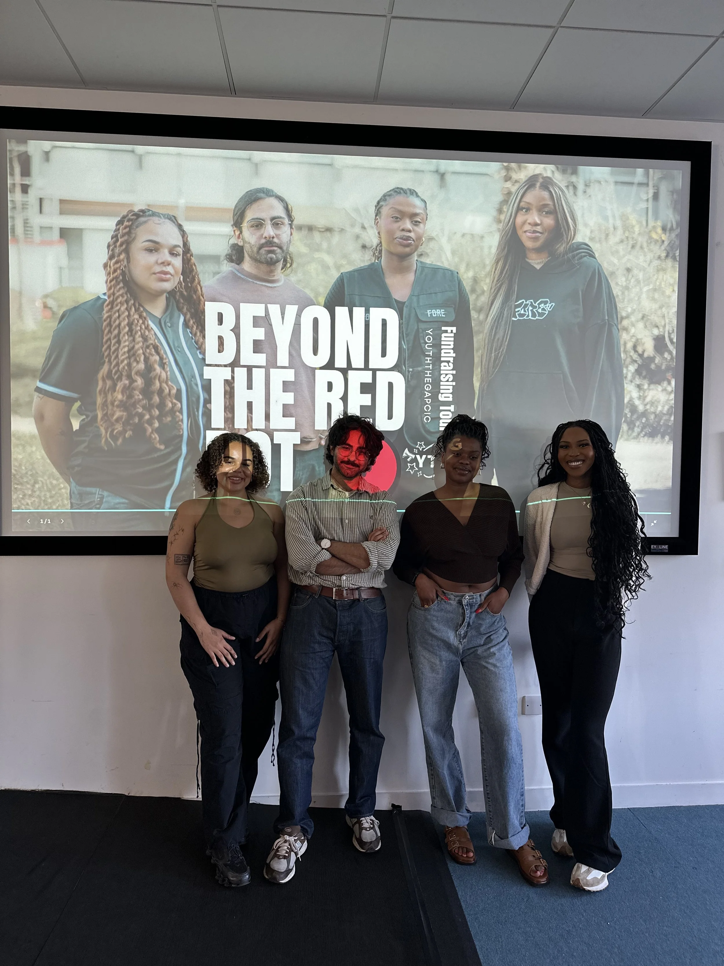 Four people standing in front of a large screen displaying a photo of four young individuals outdoors, with the text 'BEYOND THE RED' and 'Fostering Youth' on the screen.