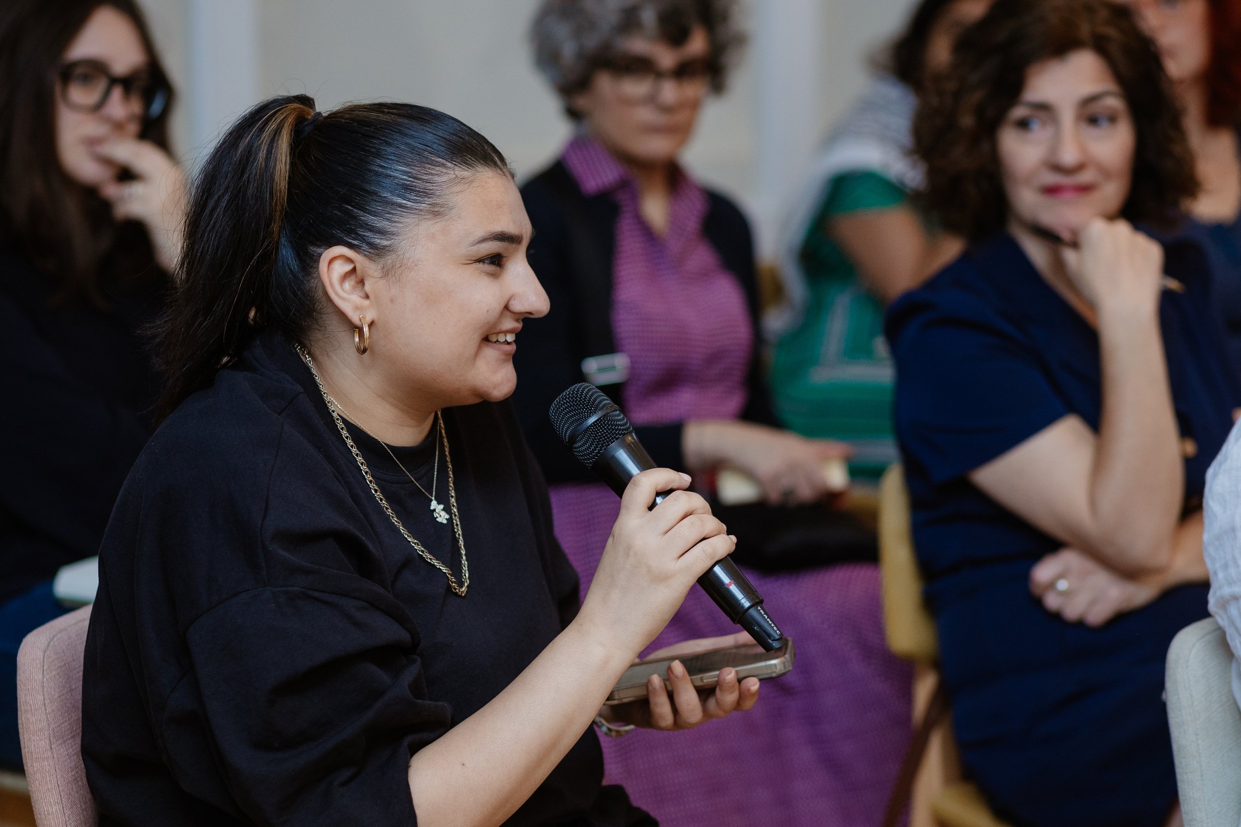 Young woman with dark hair tied back, wearing gold hoop earrings and layered necklaces, holding a microphone and smiling during a discussion in a group setting.