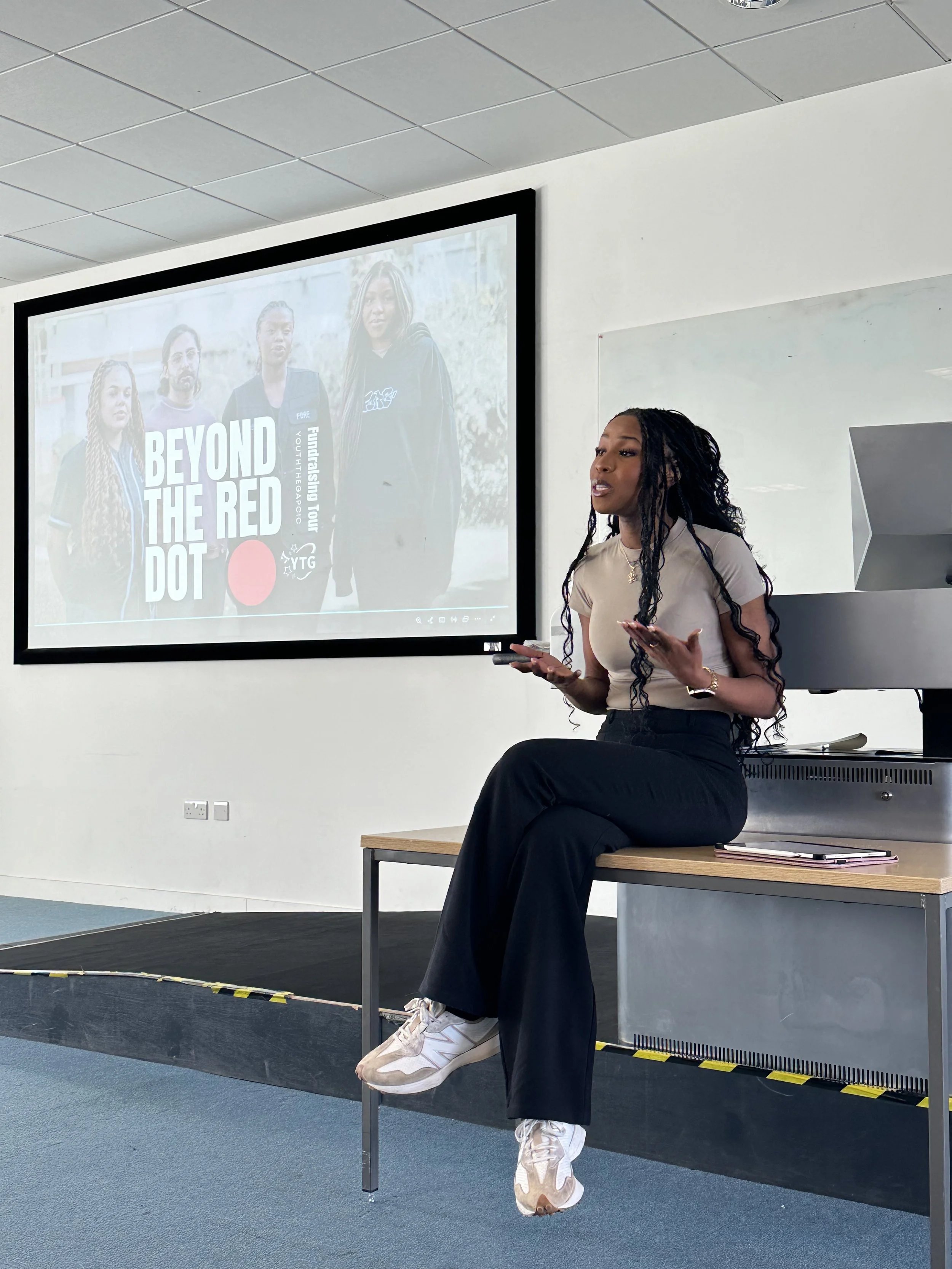 A woman with long curly black hair, wearing a beige top and black pants, sits on a desk in front of a whiteboard, giving a presentation about a youth tech program called 'Beyond the Red Dot'.
